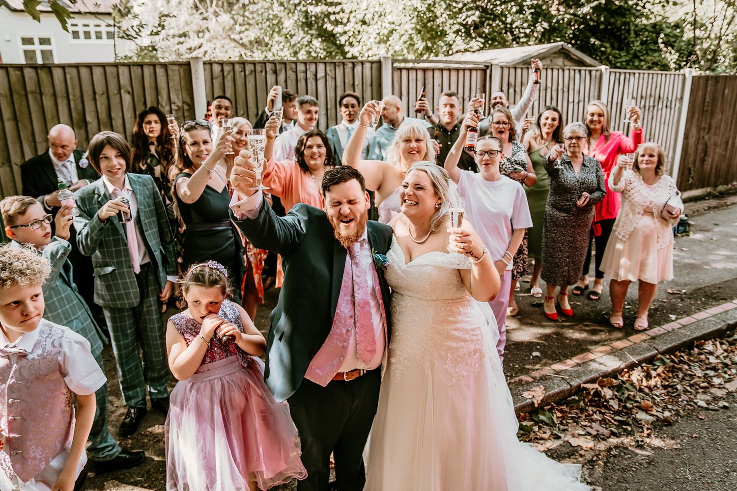 Happy bride and groom toast champagne with large group of wedding guests outdoors.