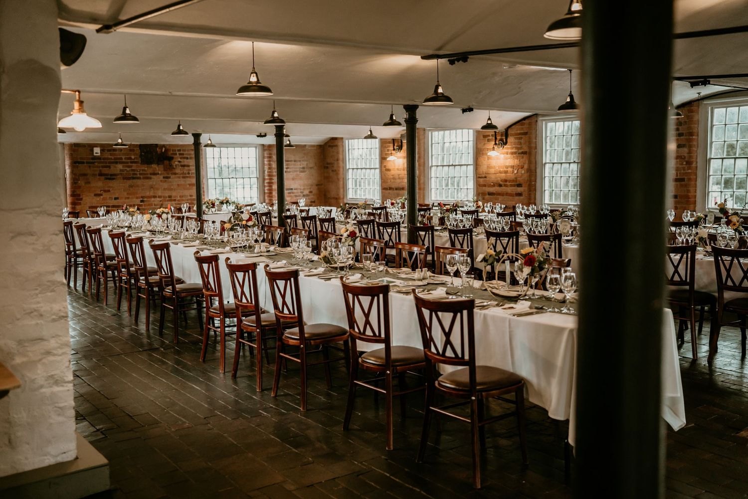 Elegant wedding reception hall with long white tables, wooden chairs, and exposed brick walls.