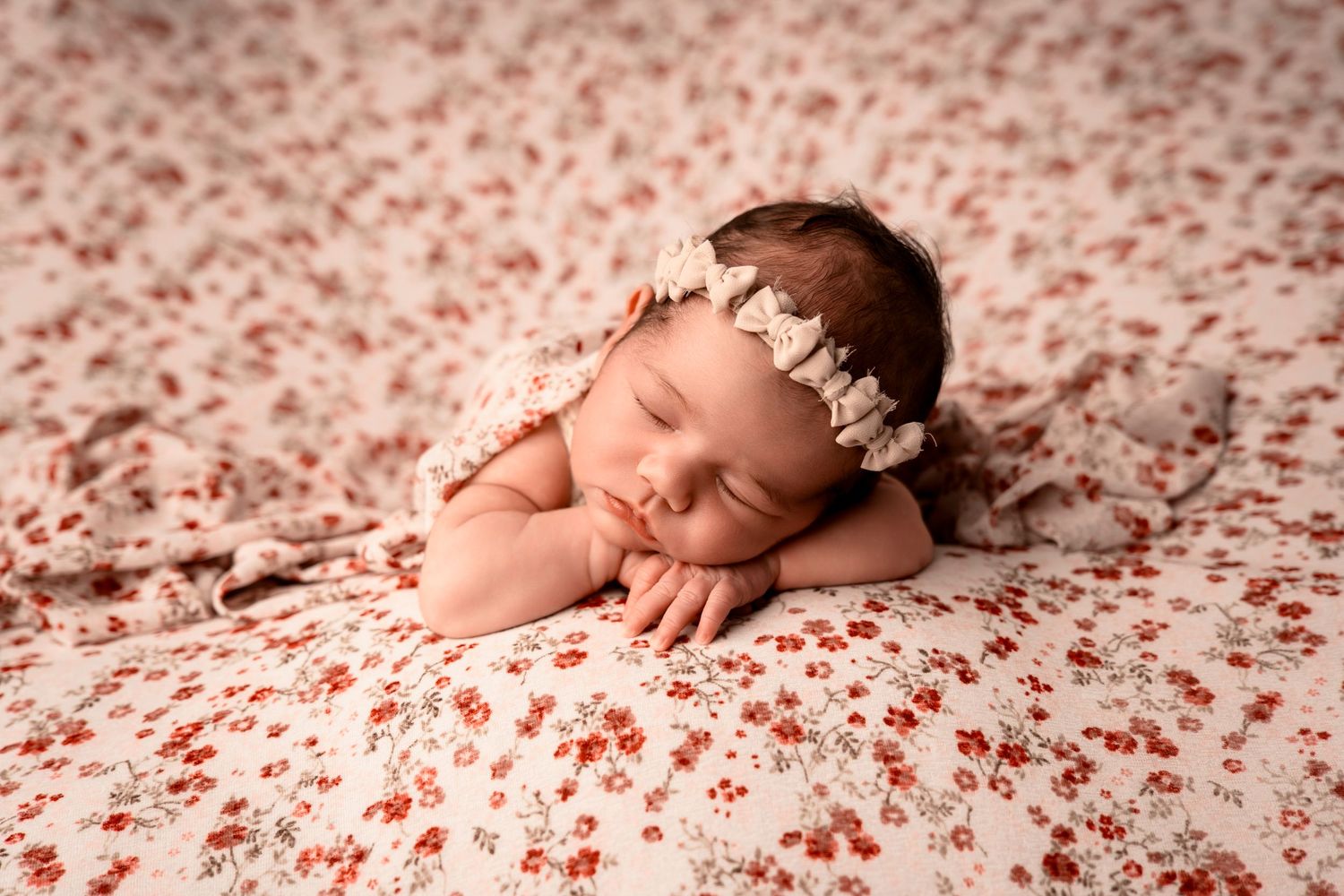 Sleeping newborn baby girl with a floral headband resting on a red and white floral blanket.