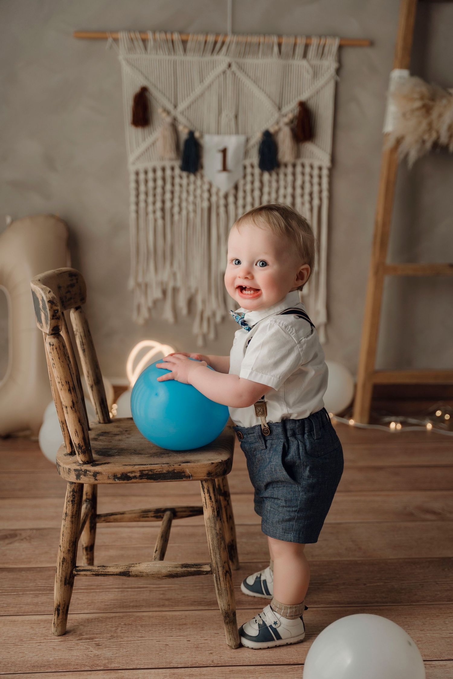 Smiling baby boy in suspenders and shorts holding a blue balloon beside a rustic wooden chair at a first birthday party.