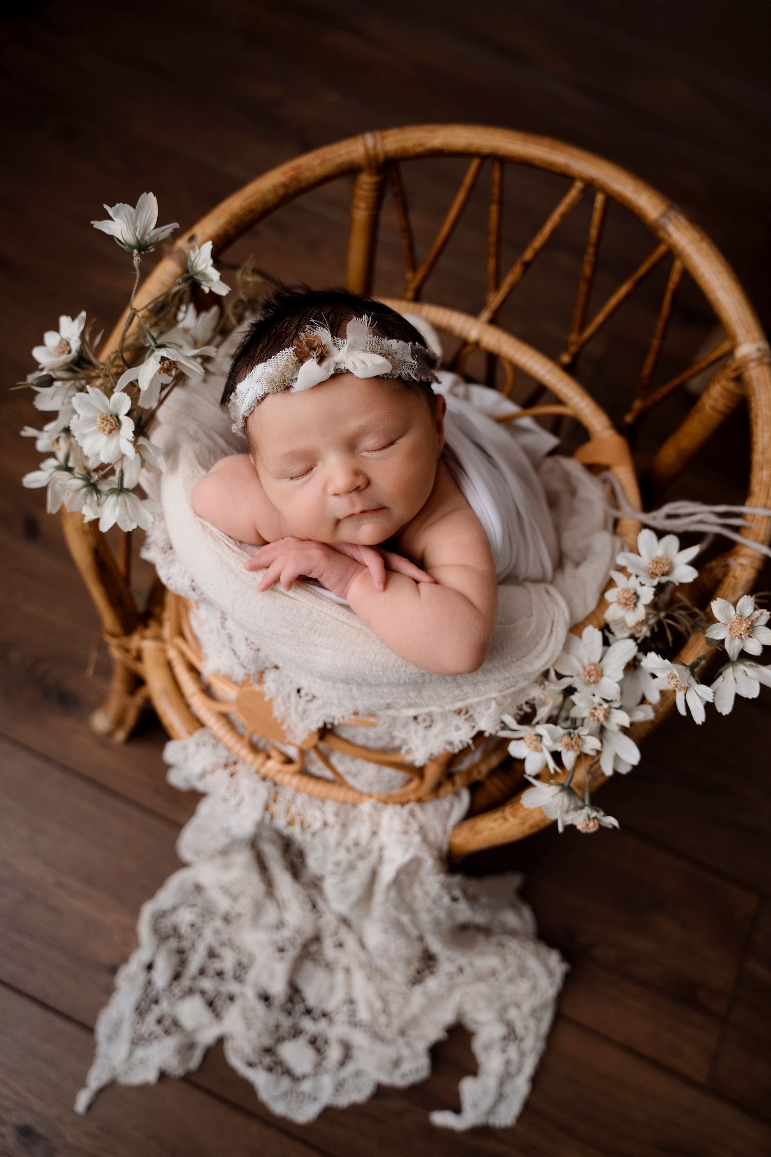 Sleeping newborn baby girl in floral headband nestled in wicker basket with white flowers.