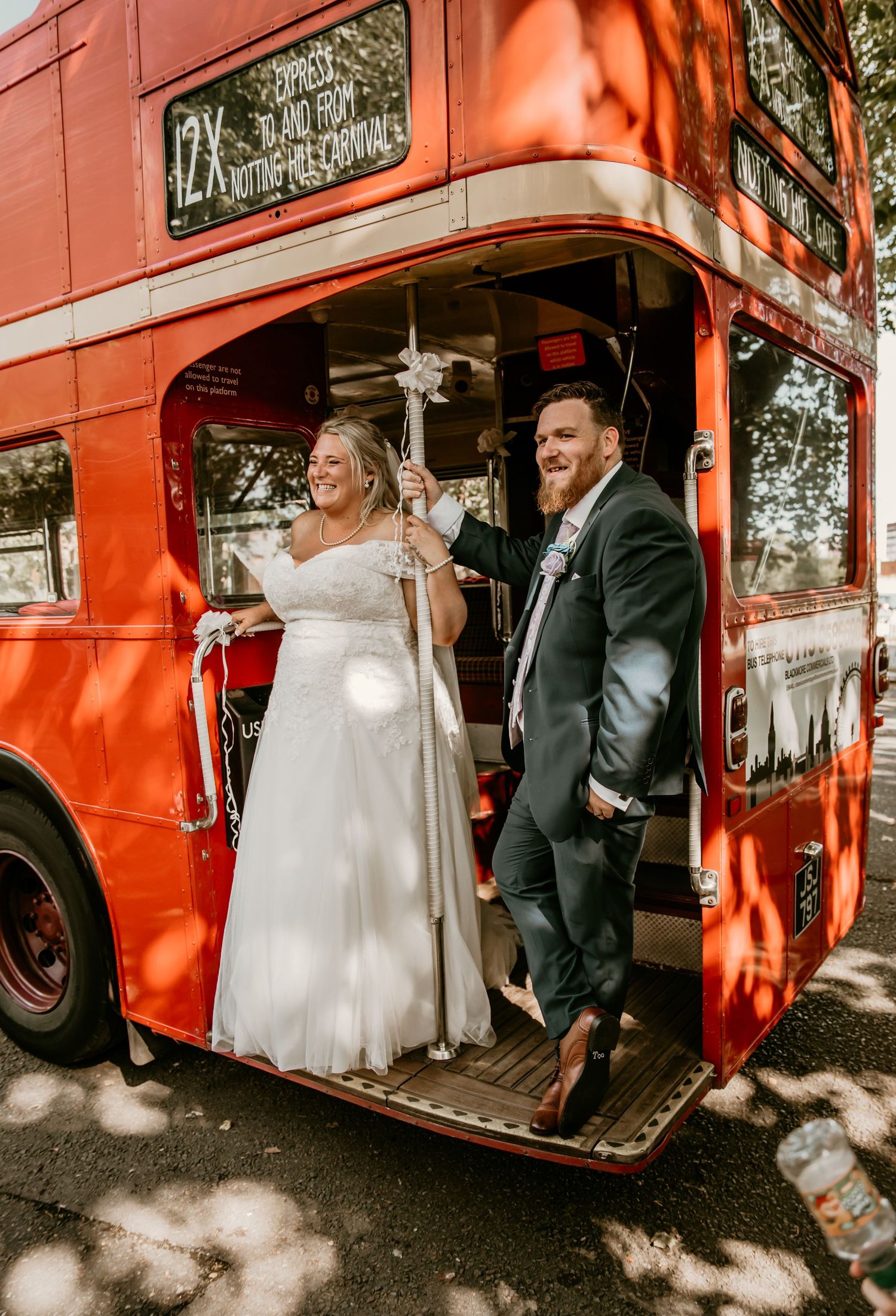 Laughing bride and groom pose on the platform of a classic red London double-decker bus.