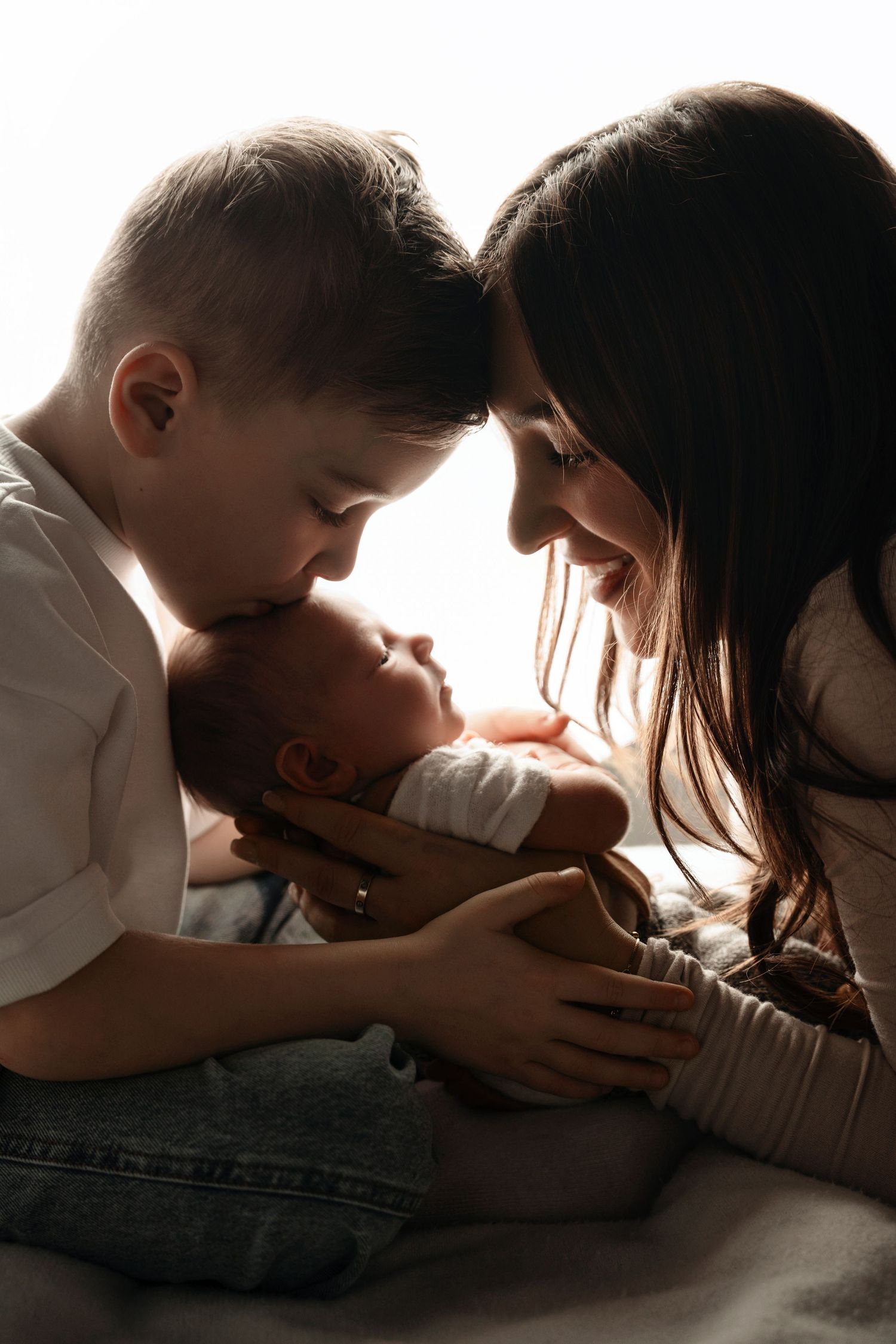 Tender moment as older sibling and mother gaze lovingly at newborn baby in warm backlit photo.