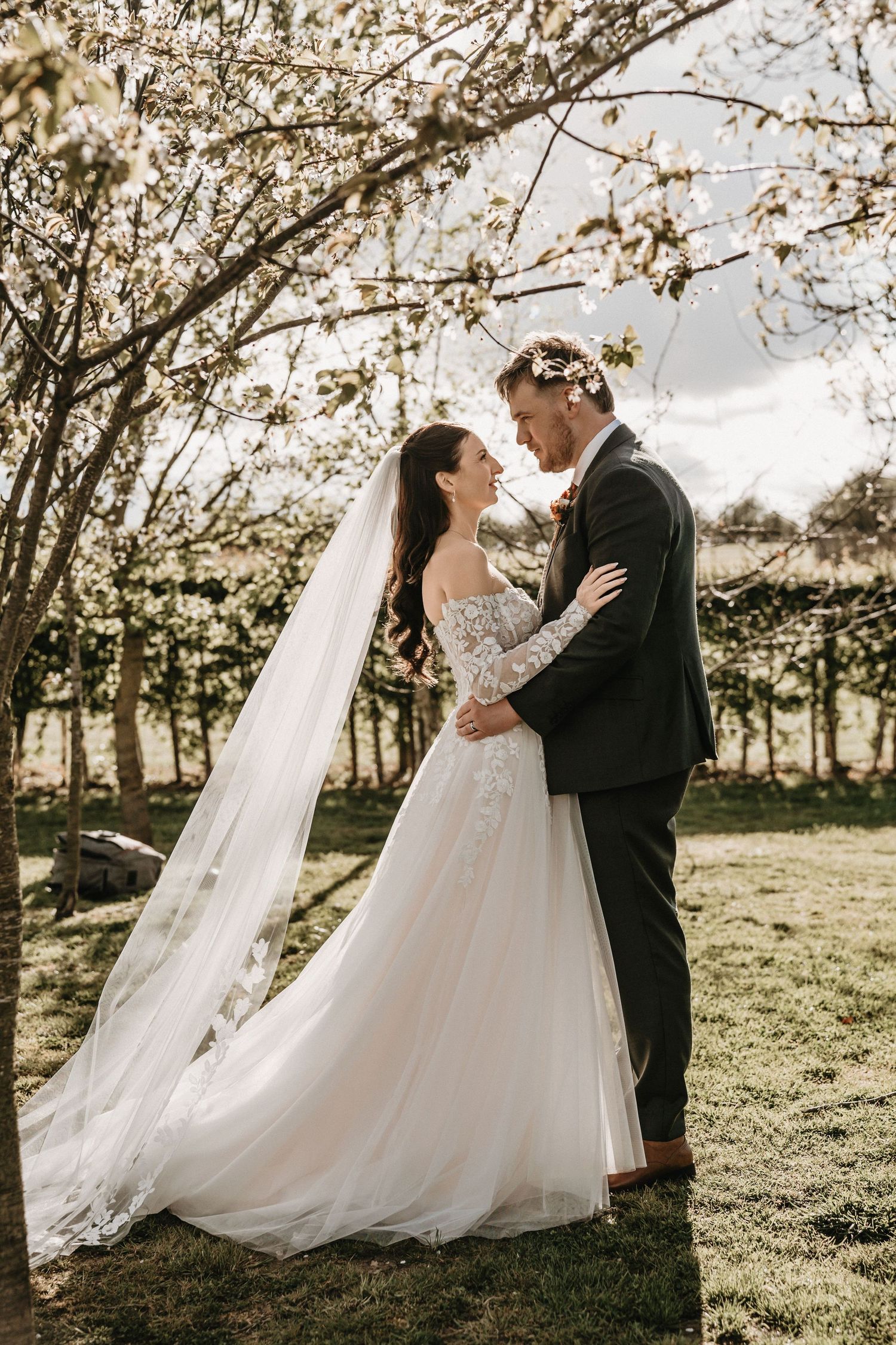 Bride in lace off-shoulder gown and groom embrace under cherry blossom trees.