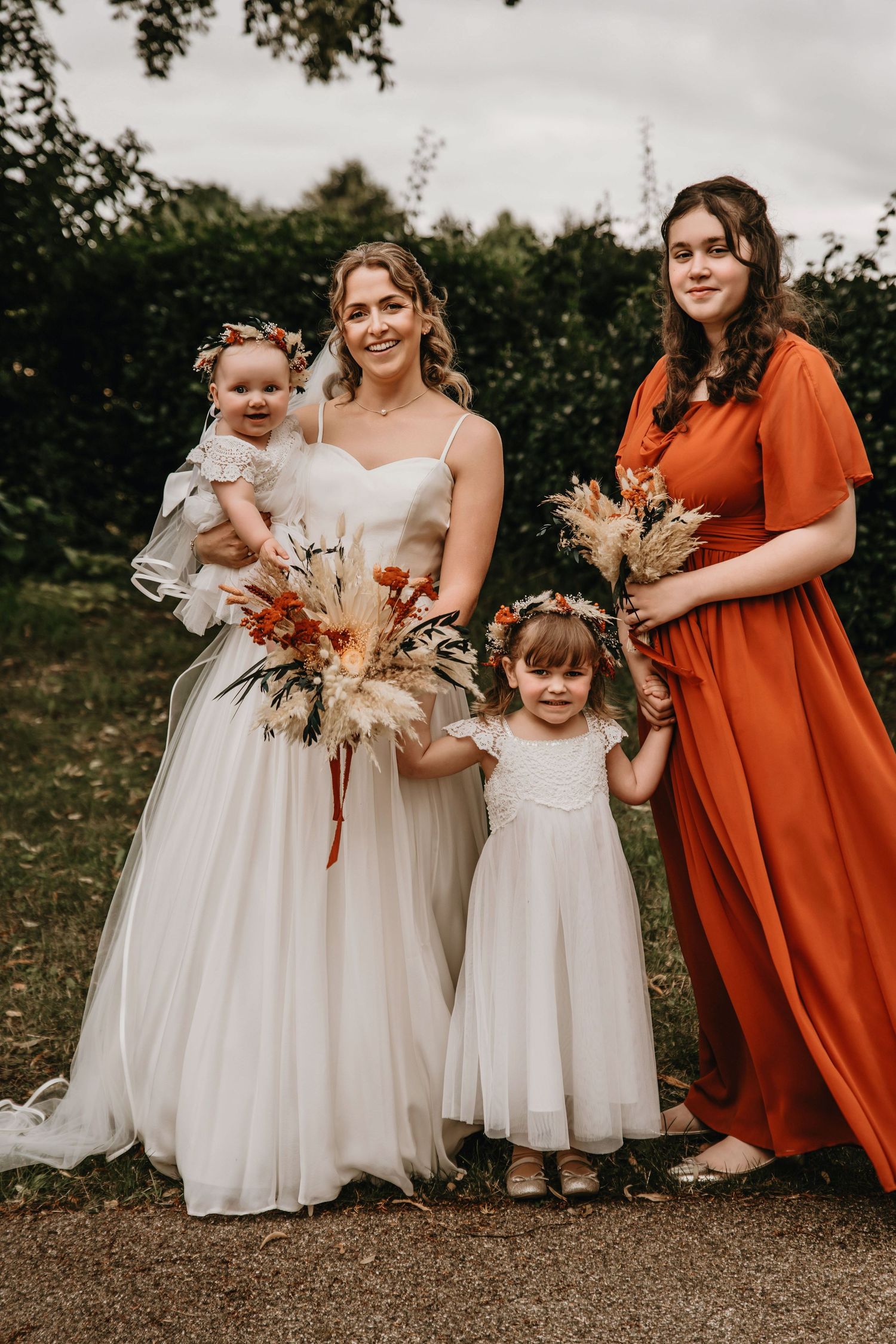 Bride in white gown with bridesmaid in orange dress and two flower girls holding bouquets outdoors.