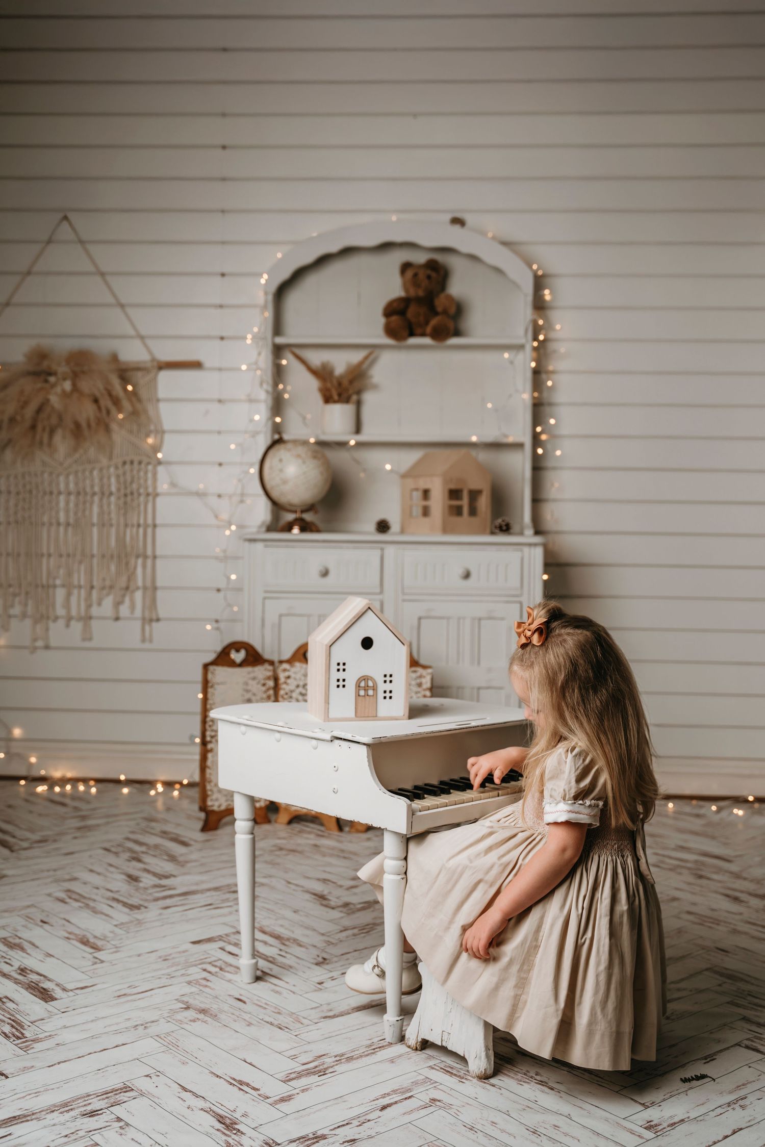 Young girl in a beige dress playing a white toy piano in a cozy decorated room with fairy lights and vintage shelving.
