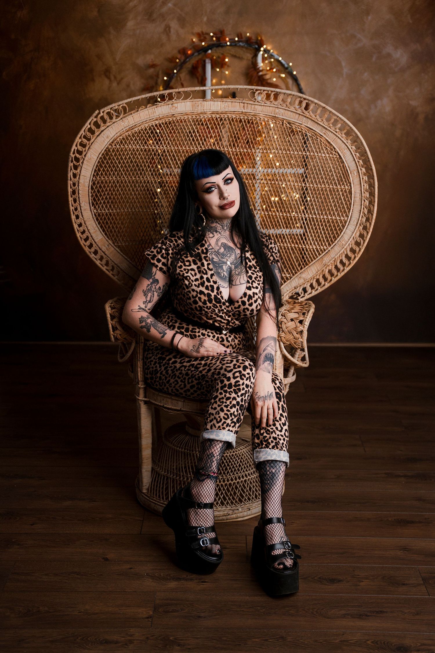 Tattooed woman in leopard print jumpsuit and platform shoes seated in a large wicker peacock chair in a dark studio.