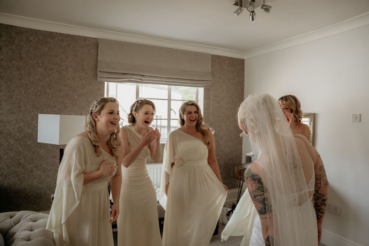 Bridesmaids in white gowns react with delight as the veiled bride reveals her wedding look indoors.