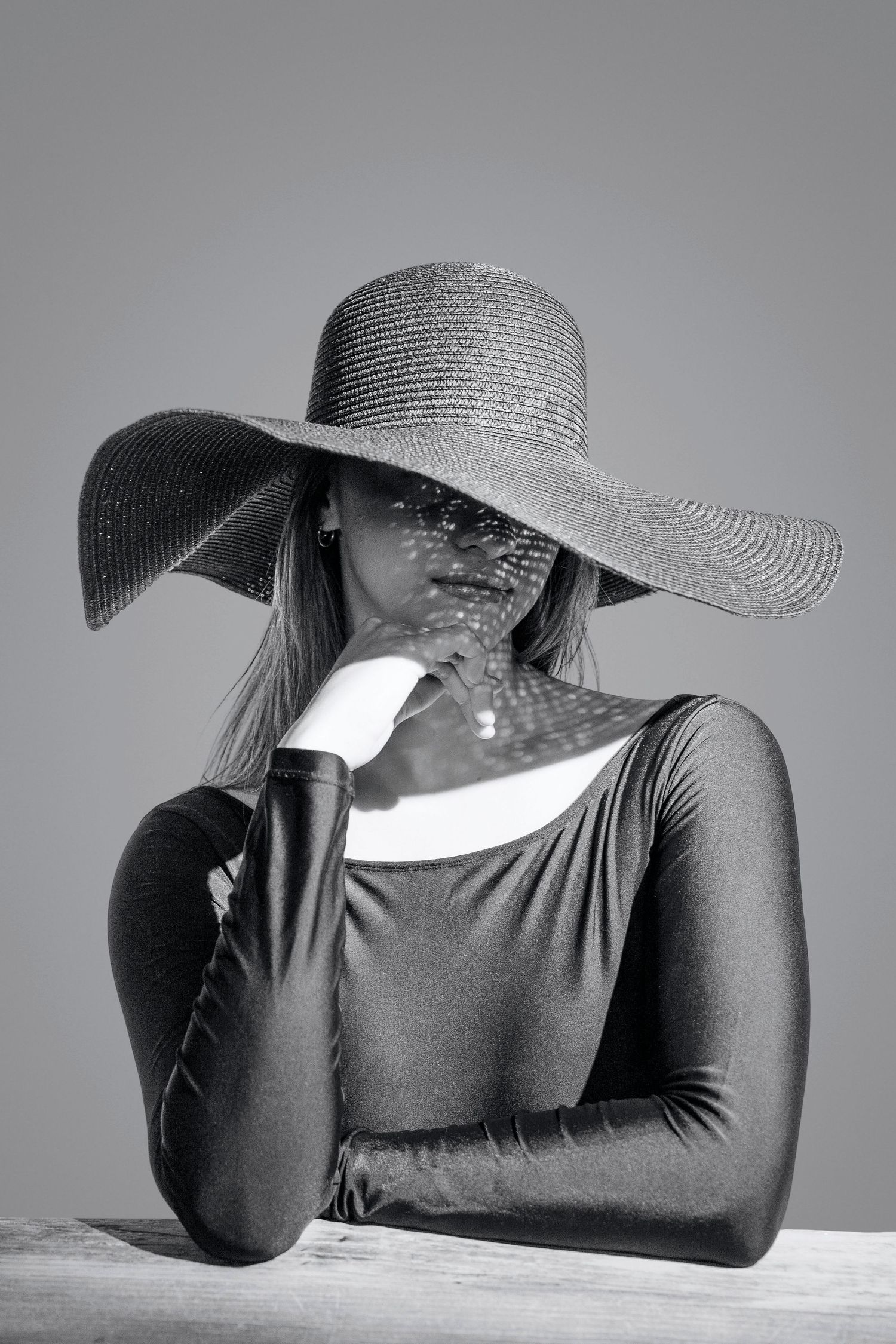 Black and white portrait of a woman in a wide-brim straw hat and fitted top, posing thoughtfully at a table.