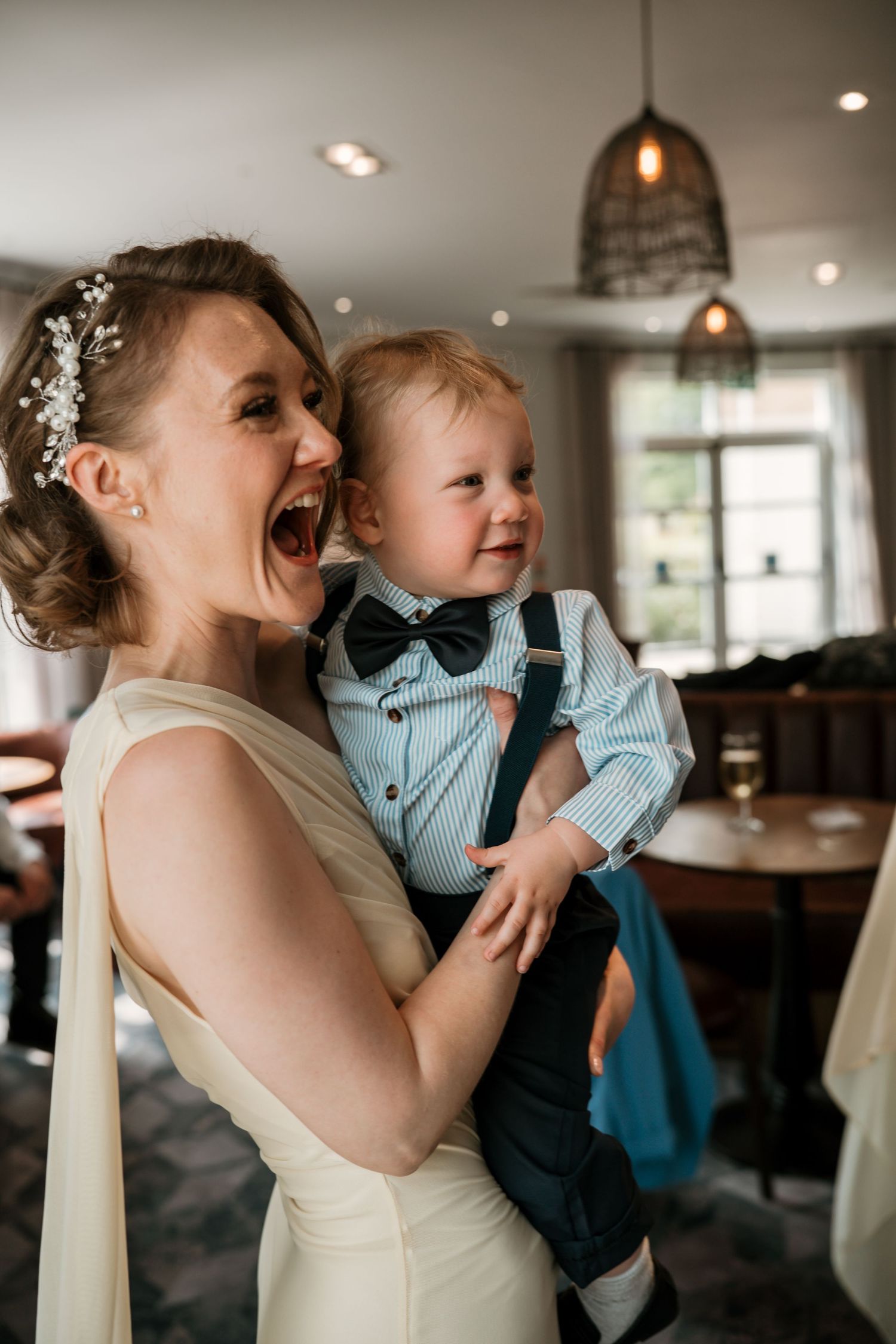 Excited bridesmaid in white dress holding adorable baby boy in bow tie.
