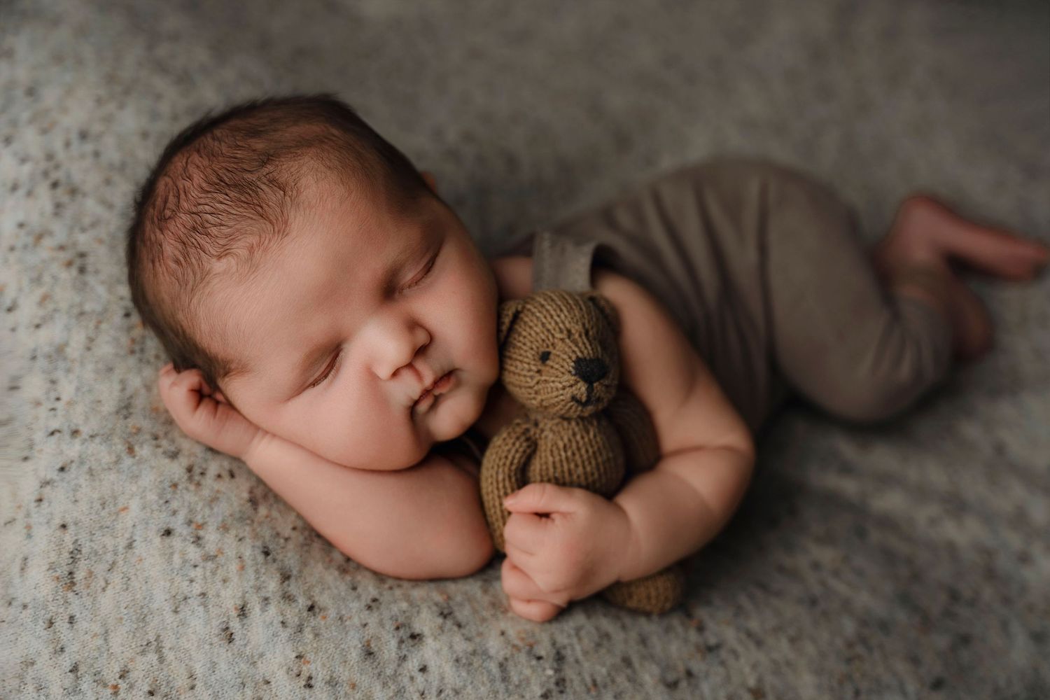 Sleeping newborn baby in gray outfit cuddling a small knitted teddy bear on a soft gray blanket.
