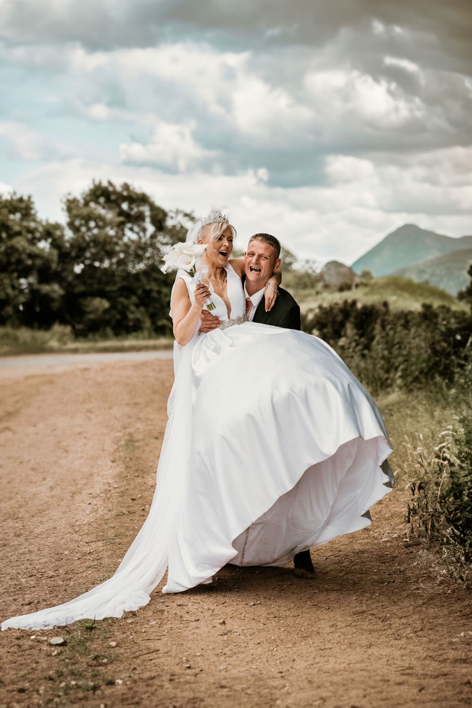 Groom lifting laughing bride in white gown on rural dirt road with mountains and trees in background.