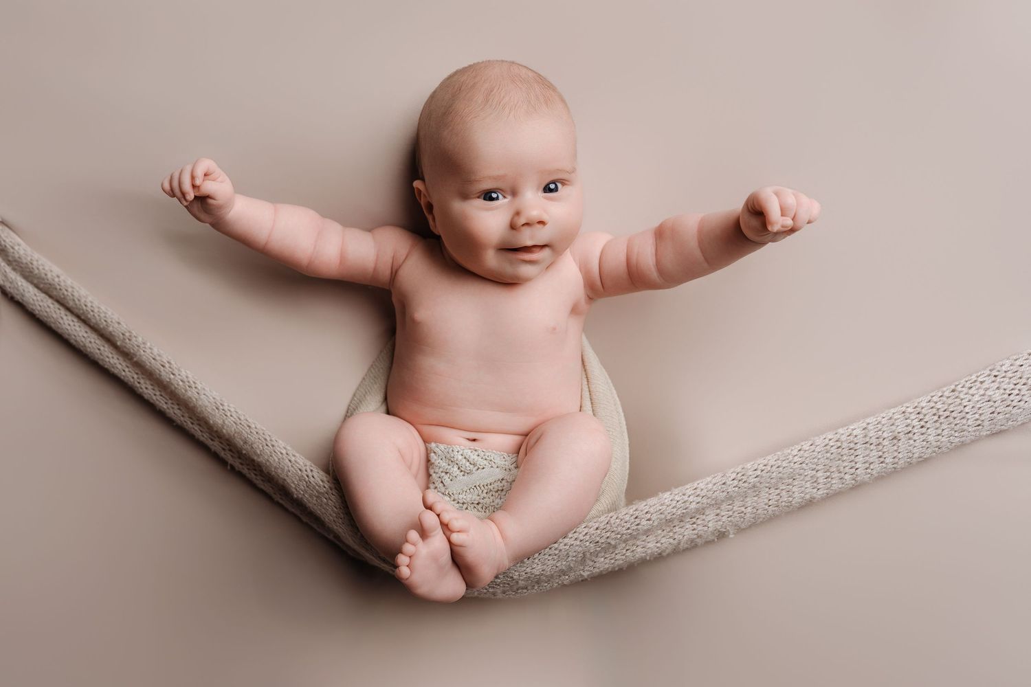 Alert baby lying on beige blanket with arms stretched wide, smiling at camera.
