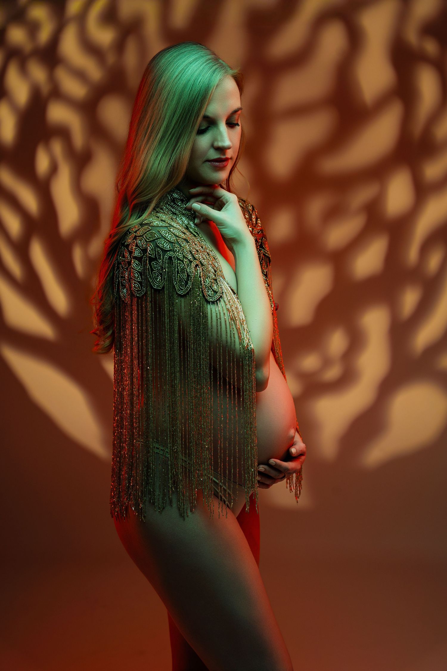 Pregnant woman in a gold fringe top posing in dramatic colored lighting with tree shadow backdrop in studio.