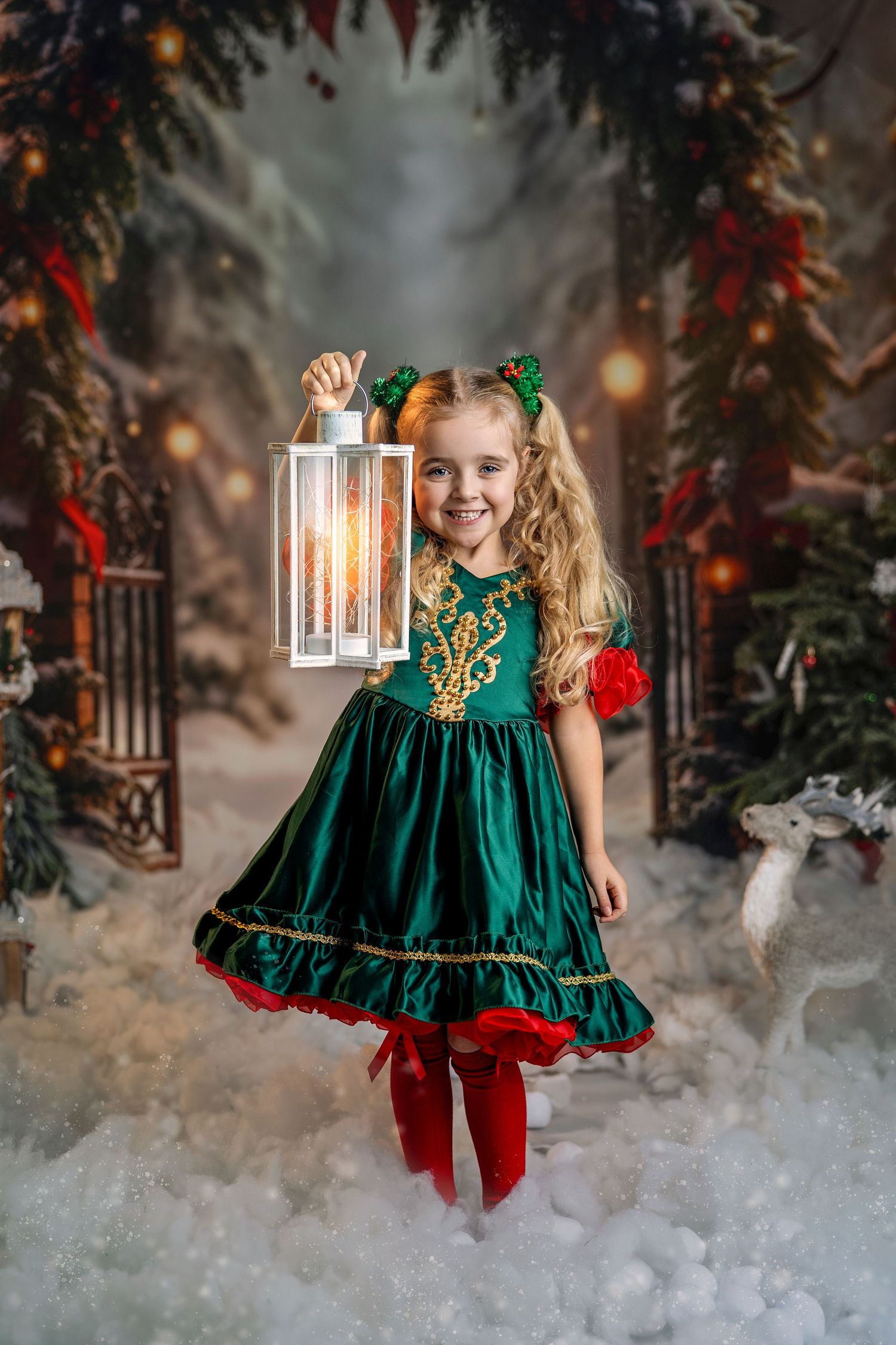 A smiling girl in a green Christmas dress holds a glowing lantern in a festive snowy holiday scene.