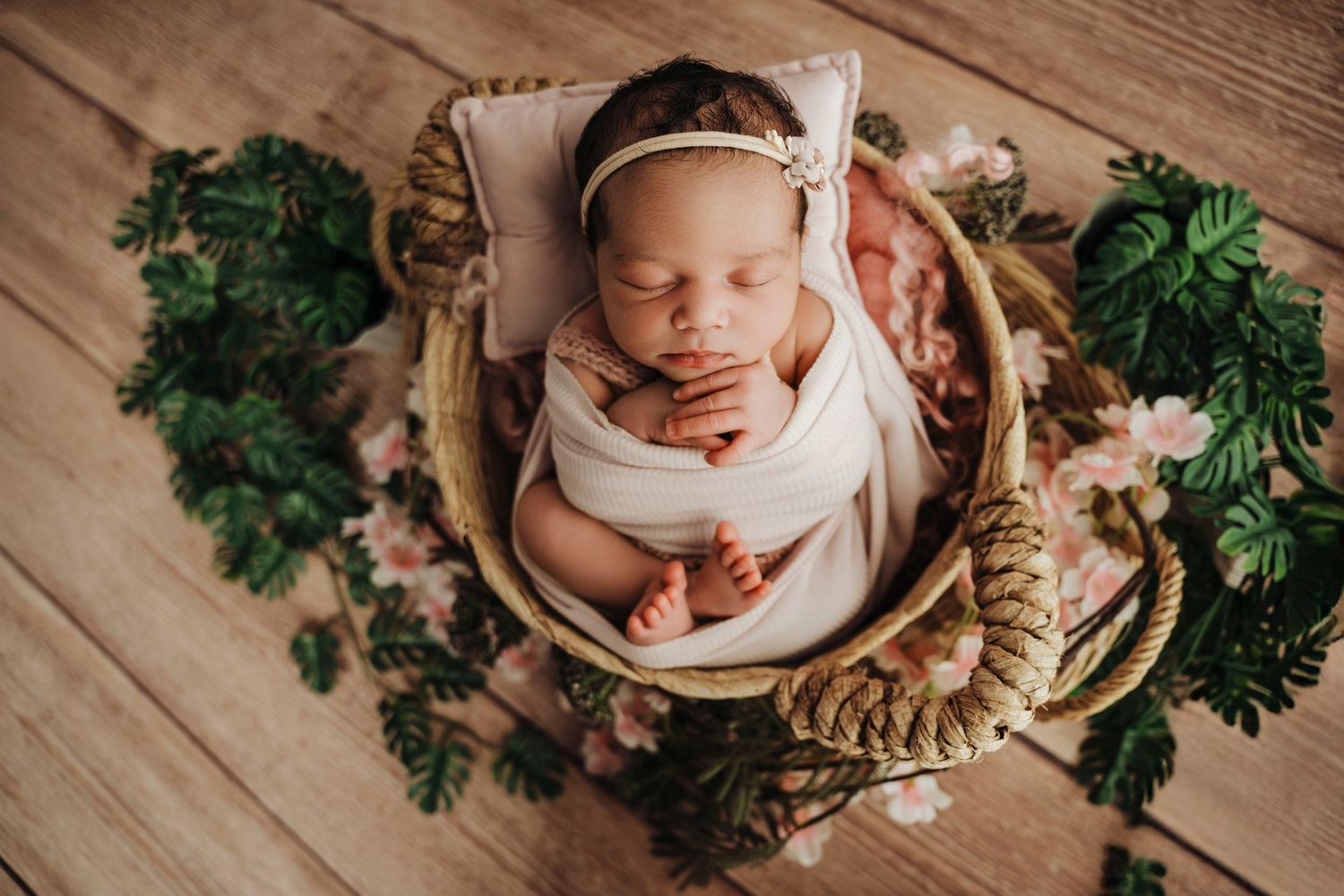 Newborn baby sleeping peacefully in a wicker basket wrapped in white cloth, surrounded by pink flowers and green leaves.