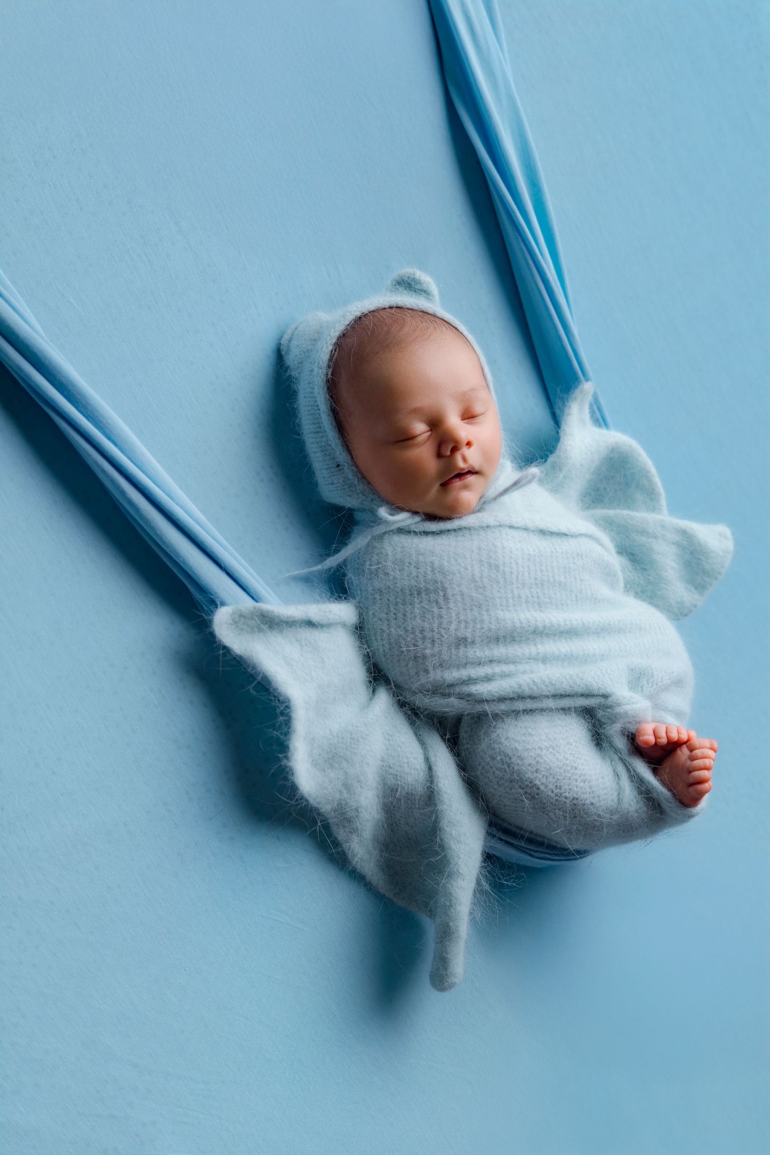 Newborn baby in light blue knit swaddle and bear bonnet sleeping on a blue fabric hammock.