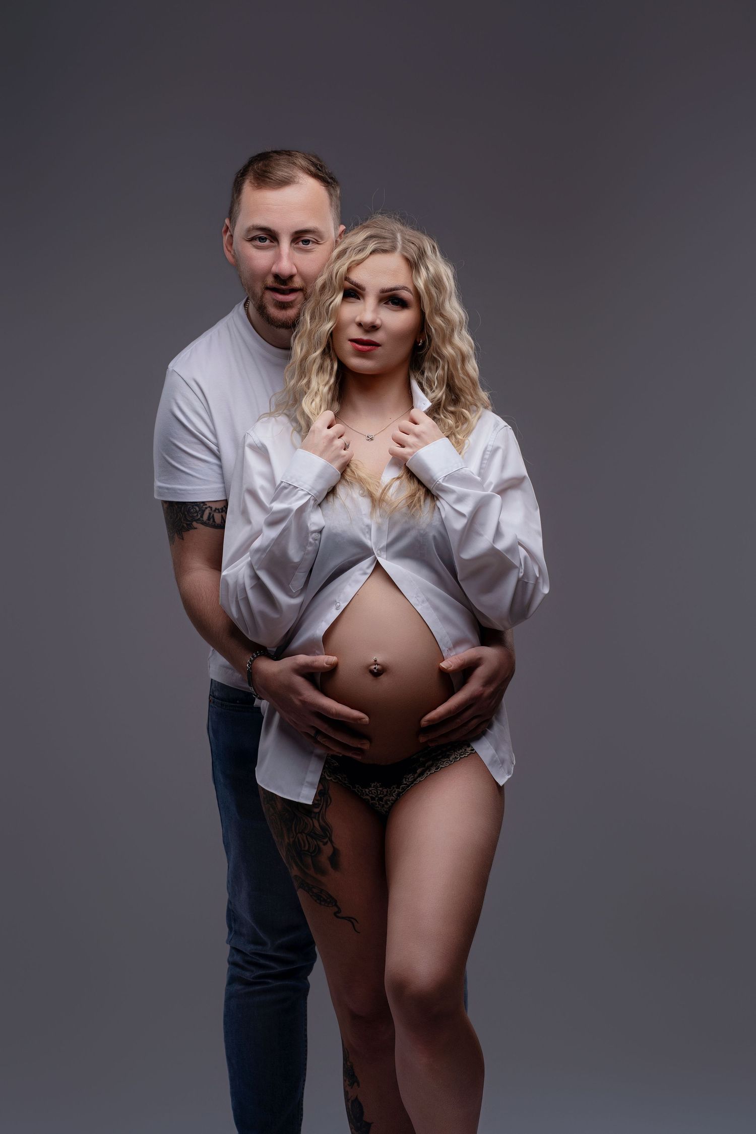 A tattooed couple poses in a studio, the man embracing his pregnant partner in a white shirt showing her baby bump.