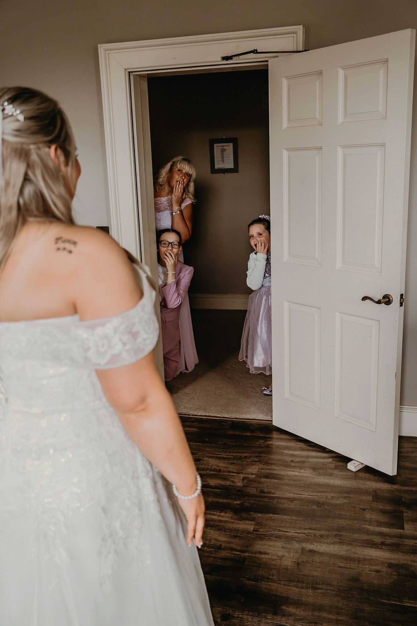 Bride in white lace off-shoulder gown surprises flower girls and bridesmaid in doorway, capturing joyful first look reactions.