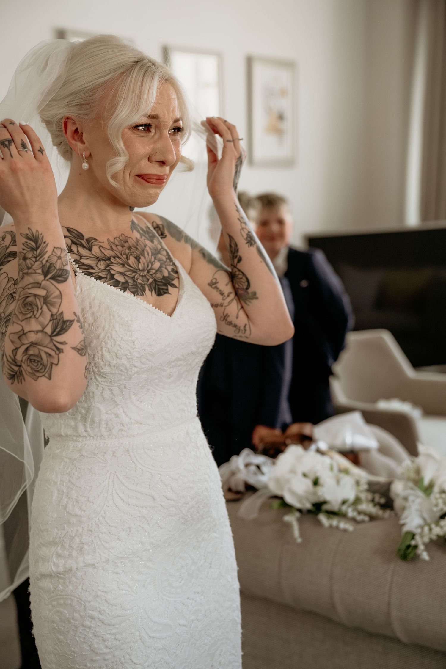 Emotional tattooed bride in lace gown wiping tears while adjusting veil.
