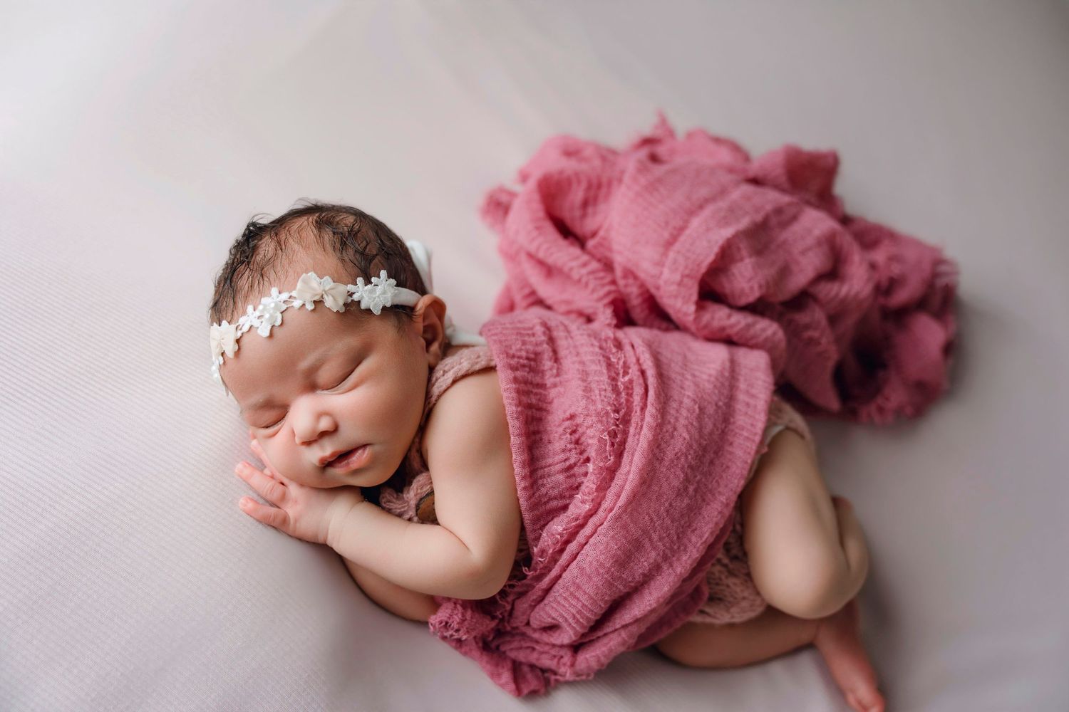 Sleeping newborn baby girl wrapped in a pink knit blanket with a white floral headband on a white background.