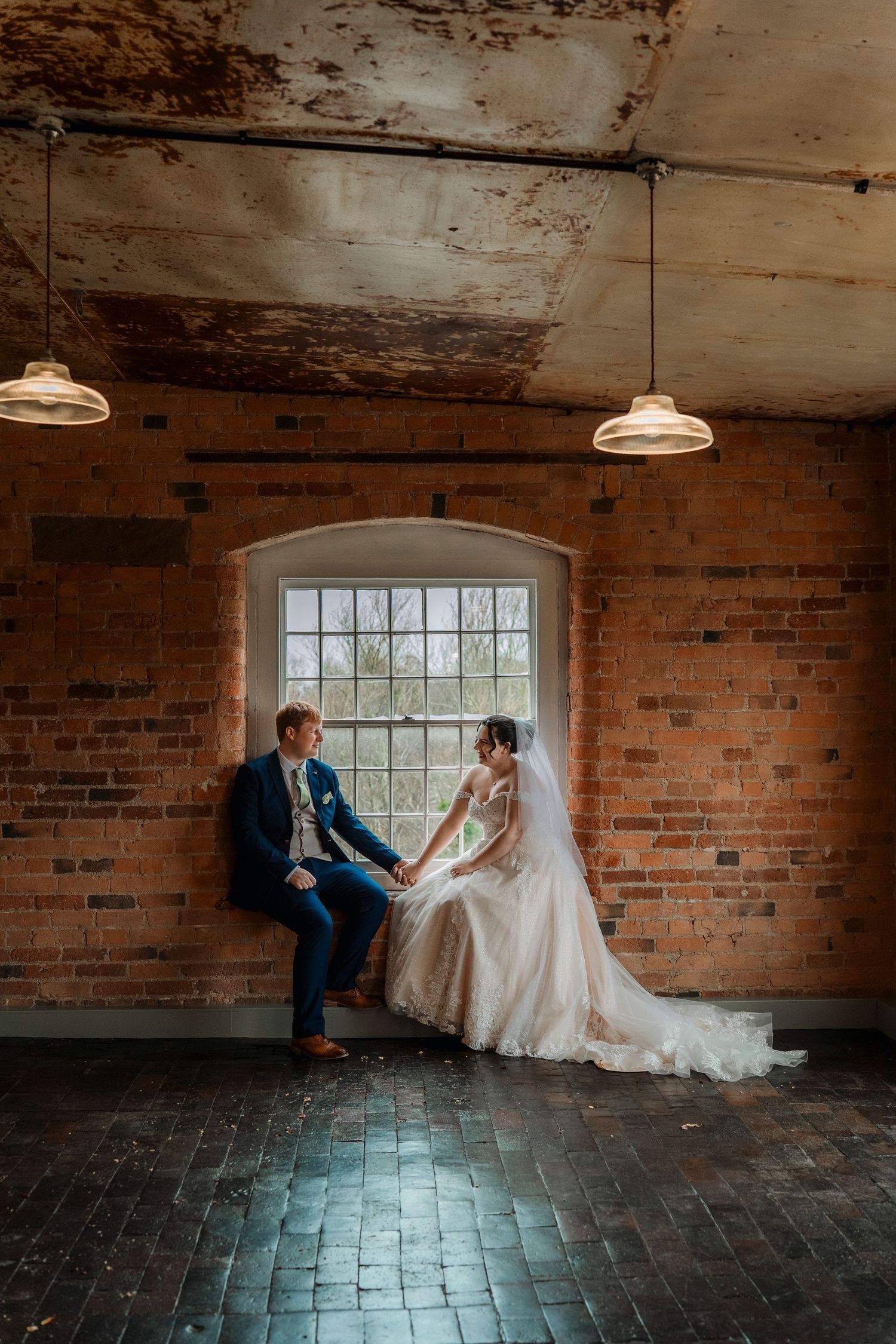 Bride and groom sitting together at arched window in rustic brick wall wedding venue with pendant lights.