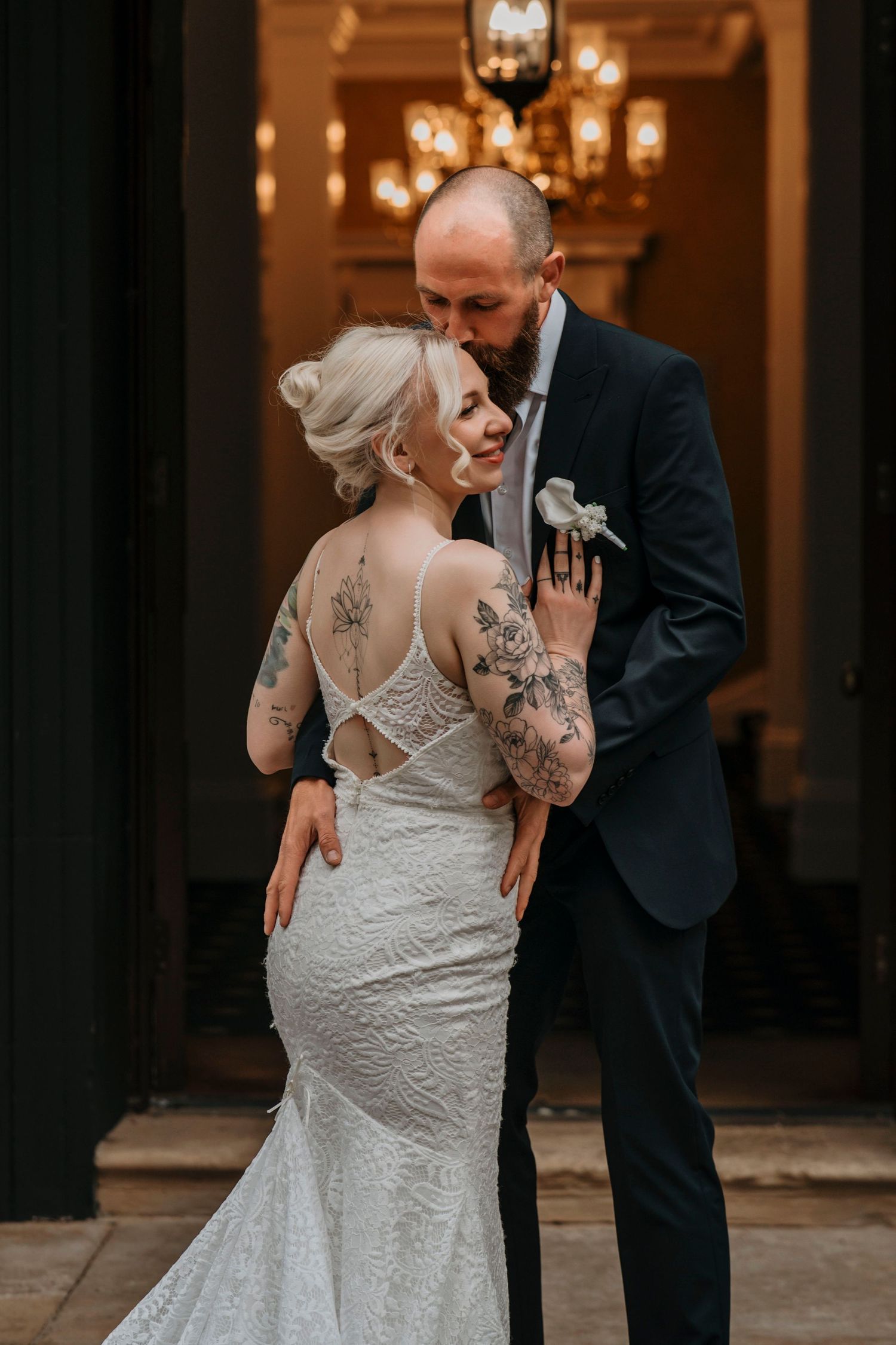 Tattooed bride in backless lace mermaid gown embraced by groom in dark suit under chandelier at wedding venue.