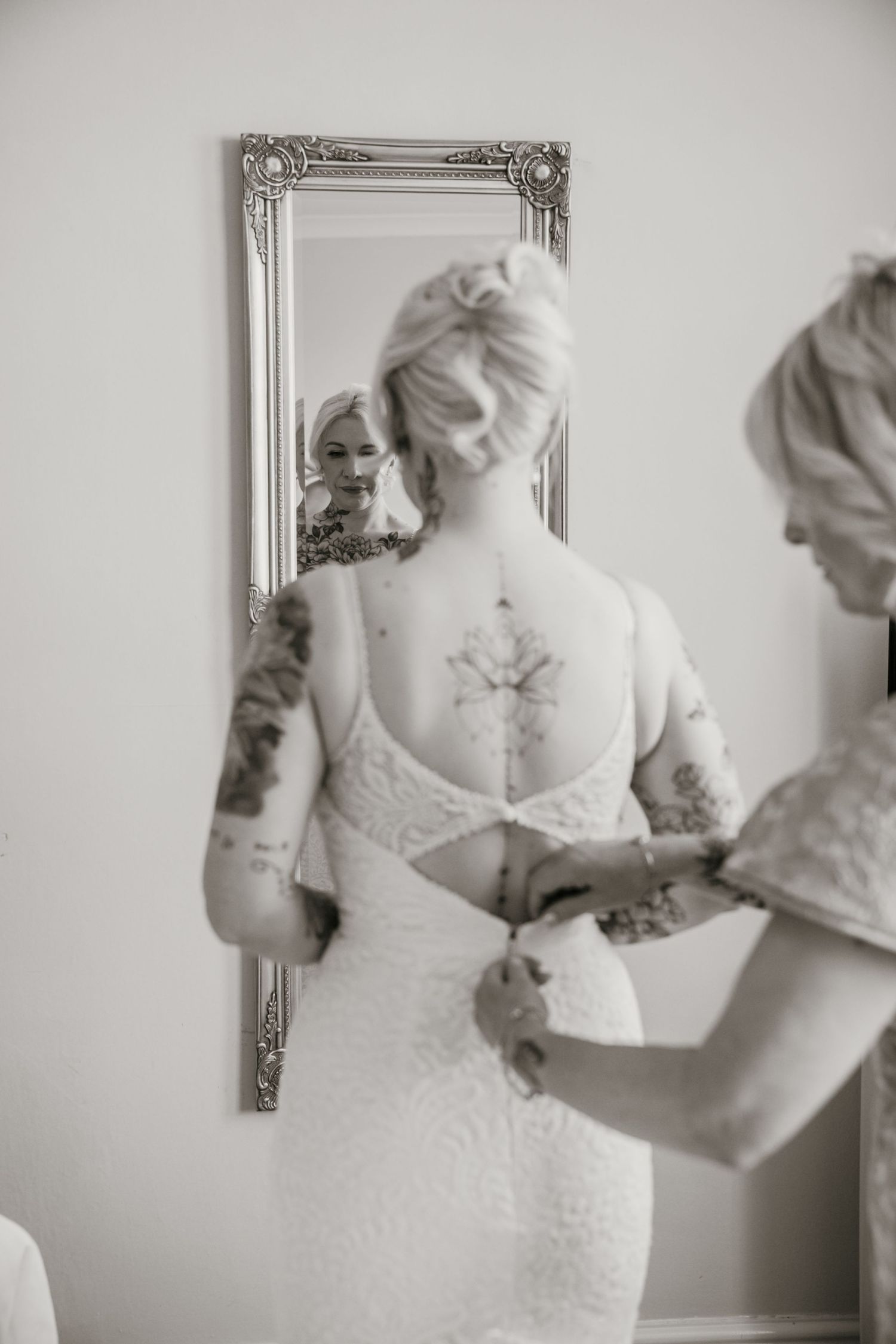 Tattooed bride in lace gown being buttoned up before ornate mirror.