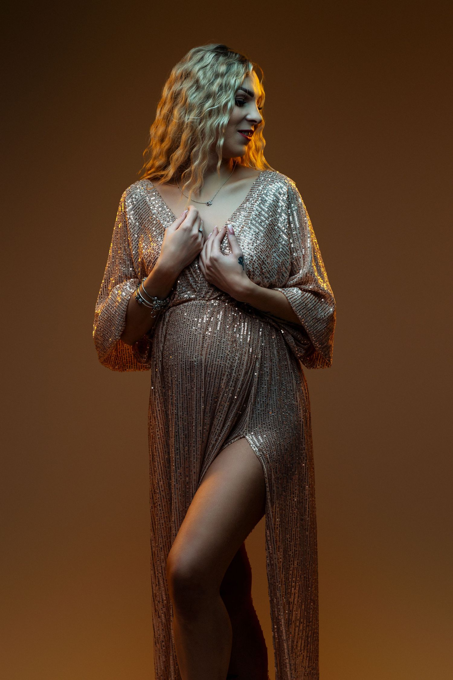 Pregnant woman in a glamorous sequined gown with a thigh-high slit, posing elegantly against a warm brown background.