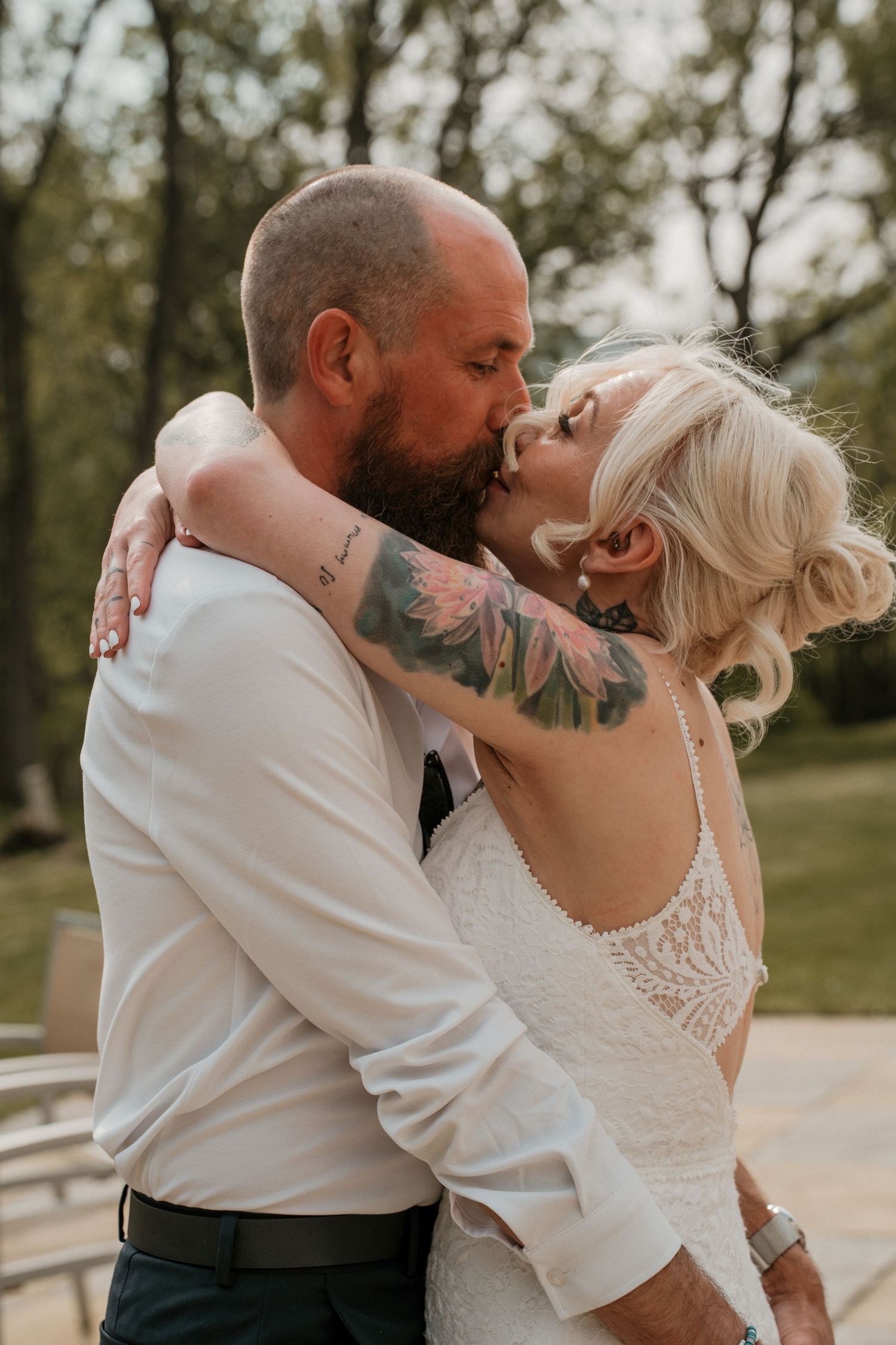 Tattooed bride and bearded groom share romantic outdoor kiss surrounded by trees after wedding.