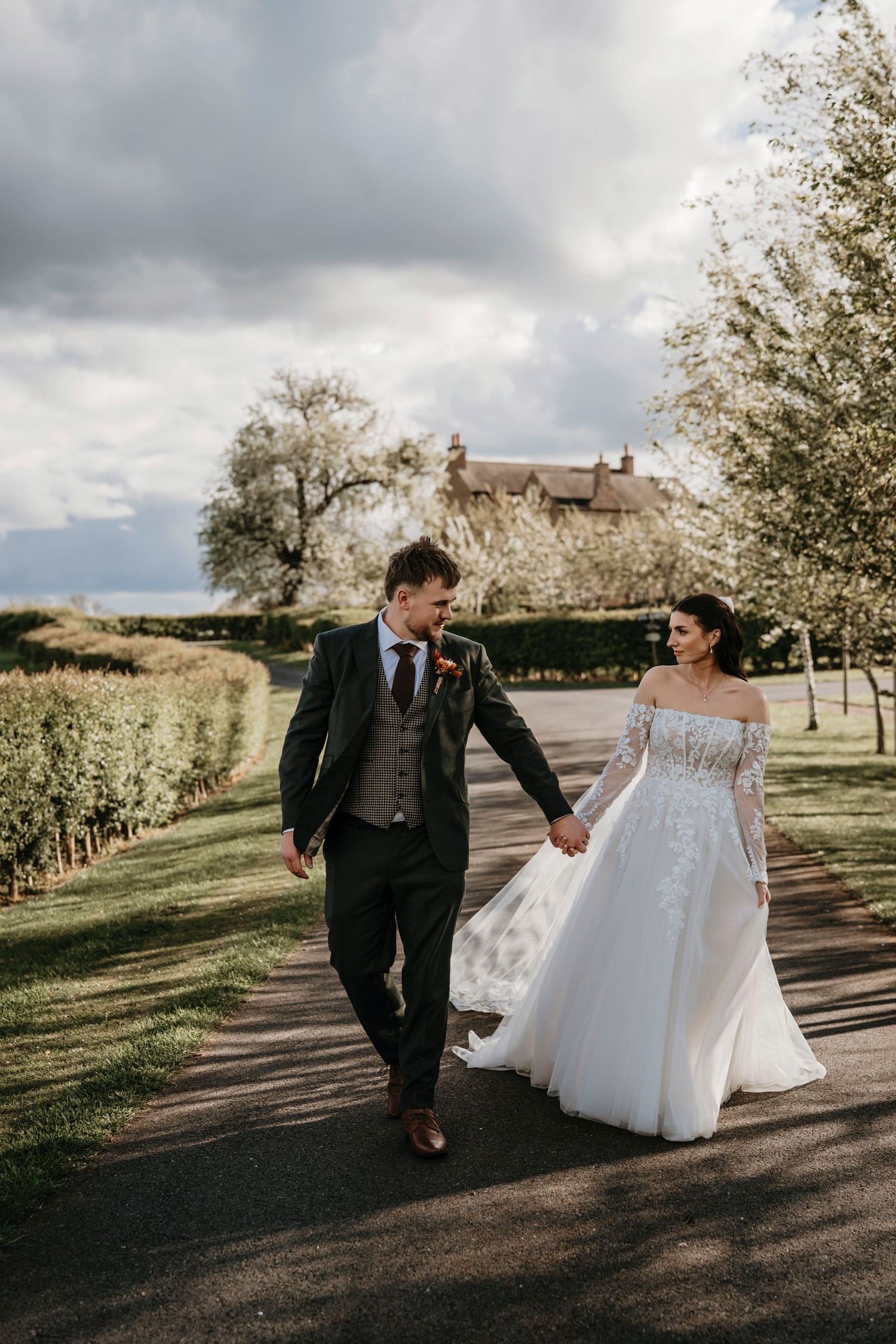 Bride and groom walk hand-in-hand along a tree-lined path, smiling at each other on their wedding day.