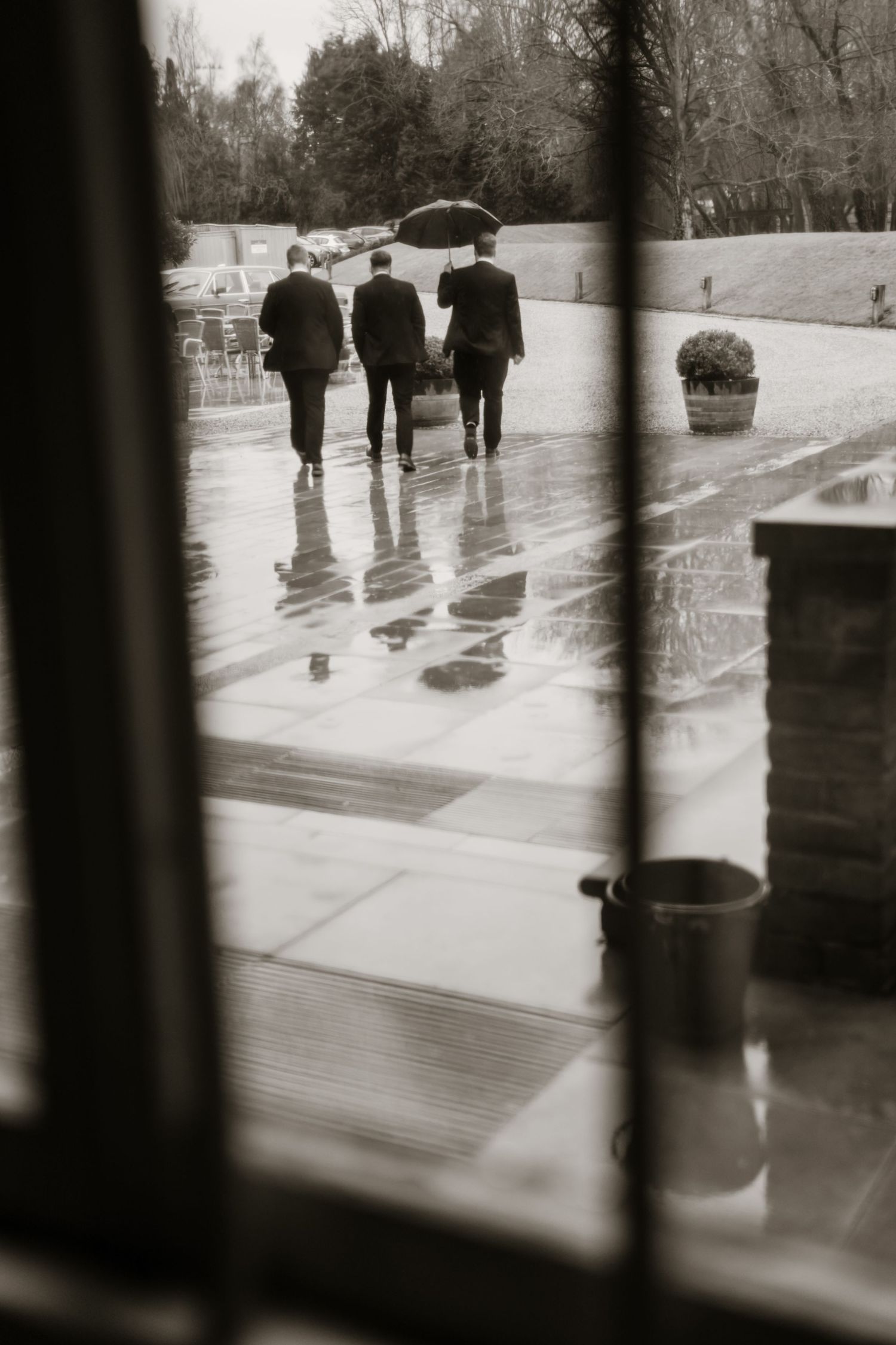 Black and white photo of three suited groomsmen walking in rain under an umbrella.