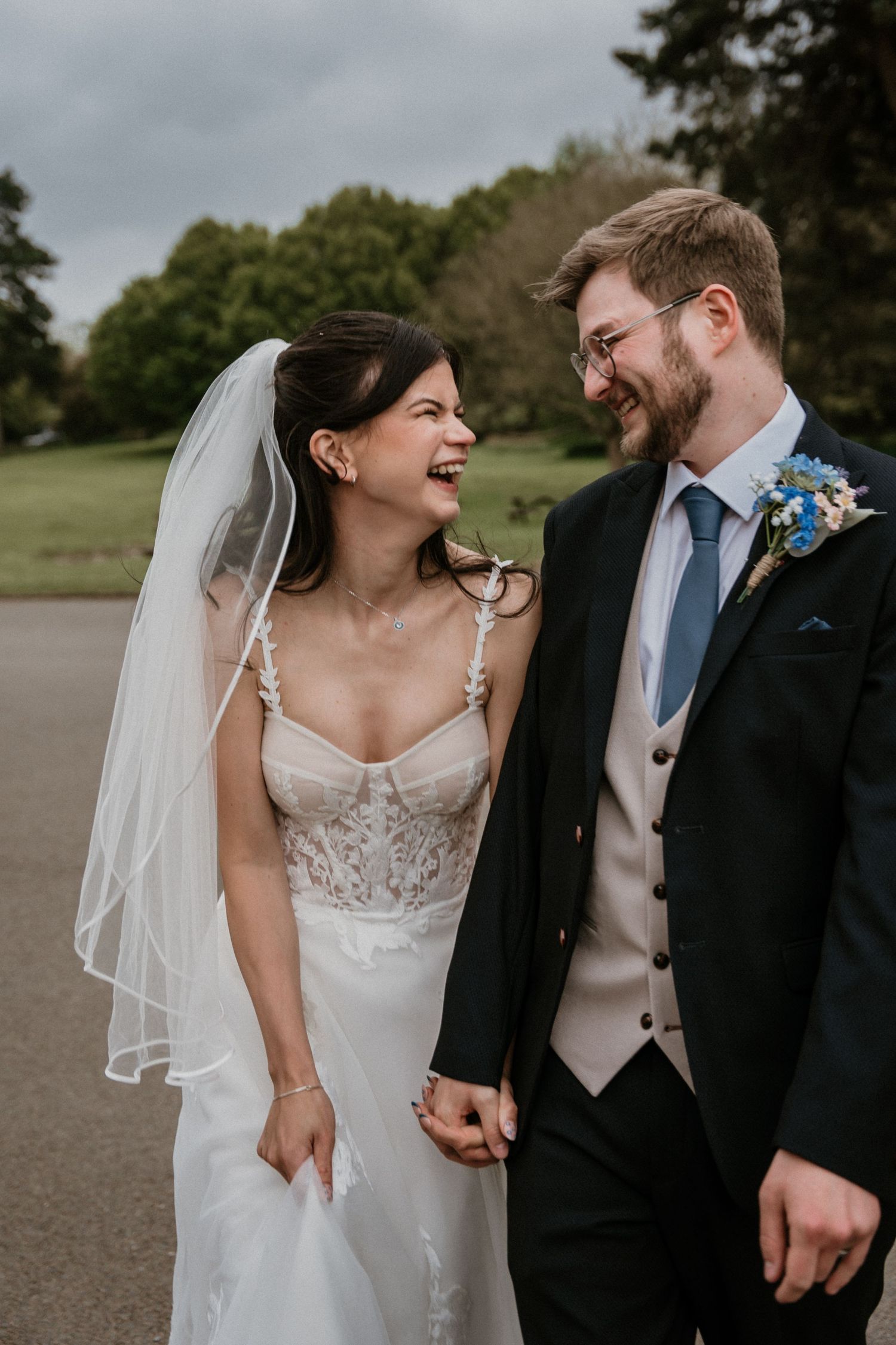 Laughing bride in lace corset gown with veil and groom in navy suit holding hands outdoors.