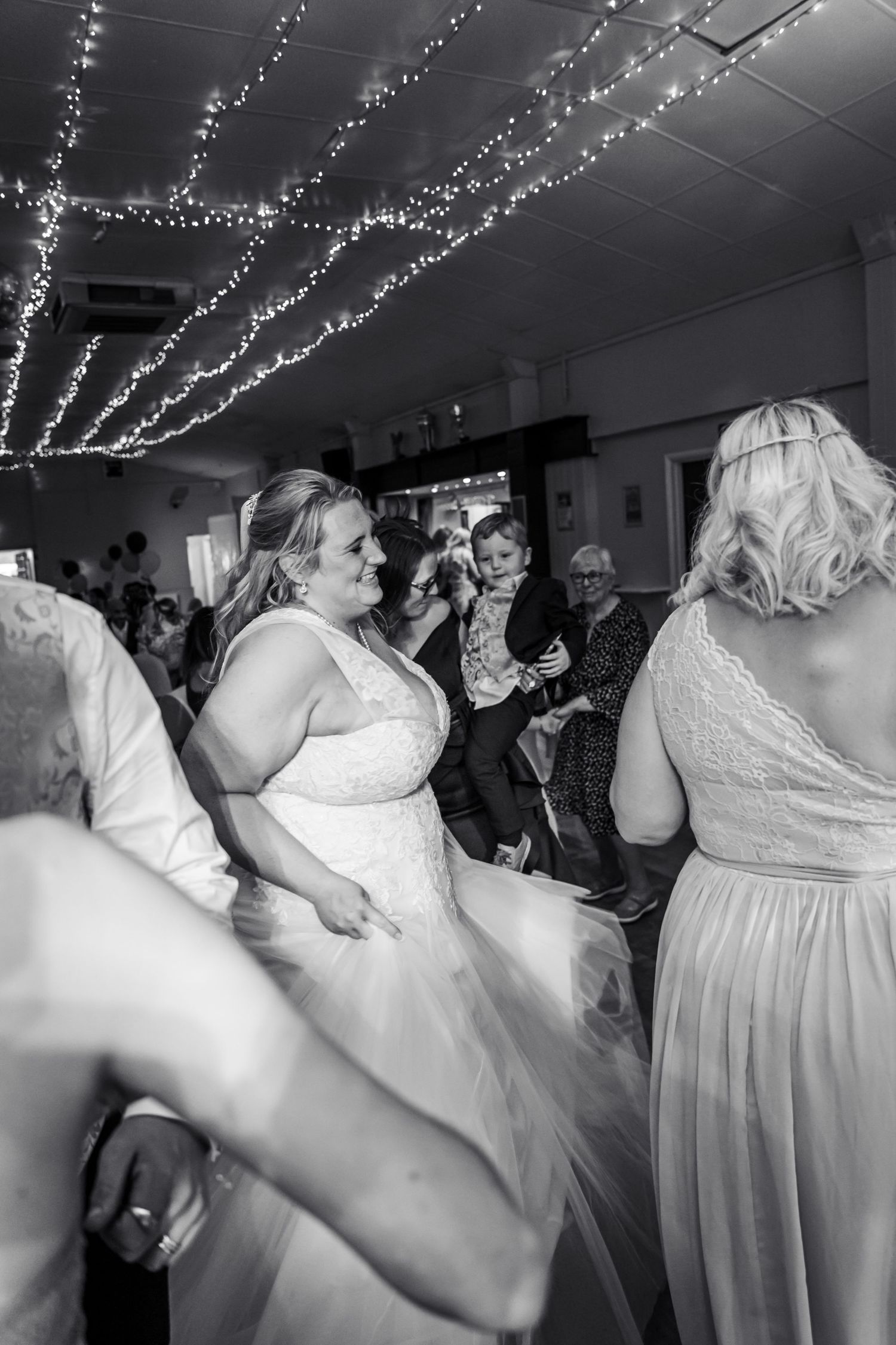 Bride in lace gown dances joyfully under fairy lights at wedding reception in black and white photo.