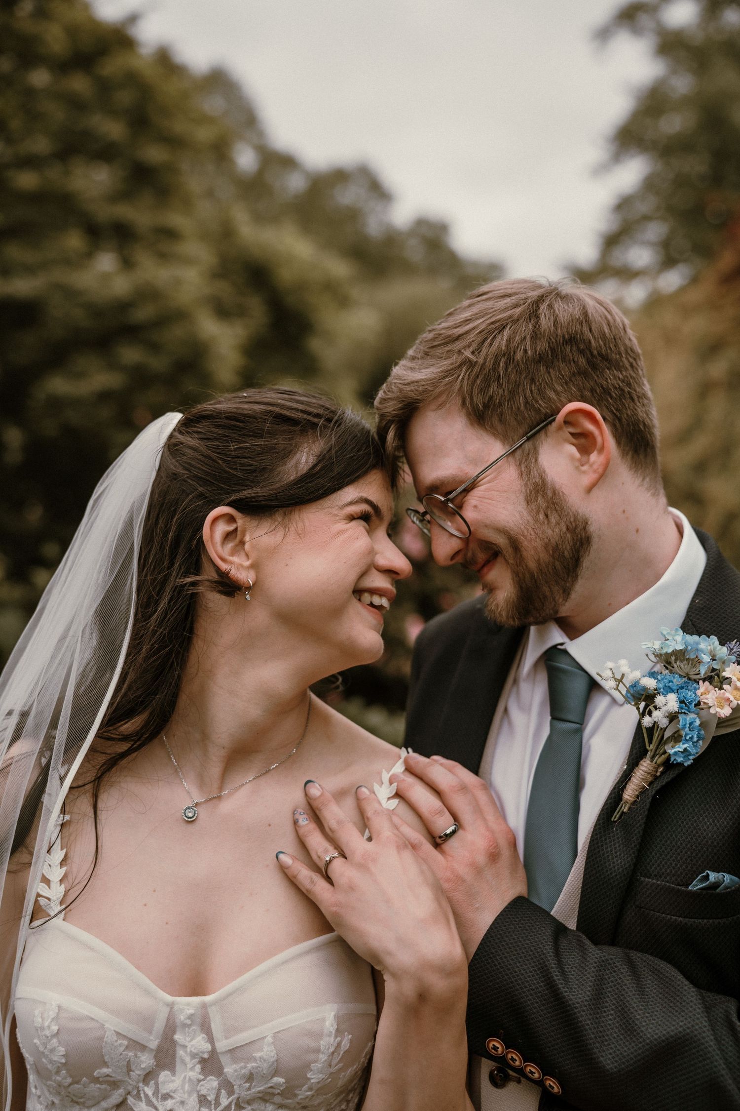 Smiling bride and groom touch foreheads intimately in a close-up wedding portrait surrounded by greenery.