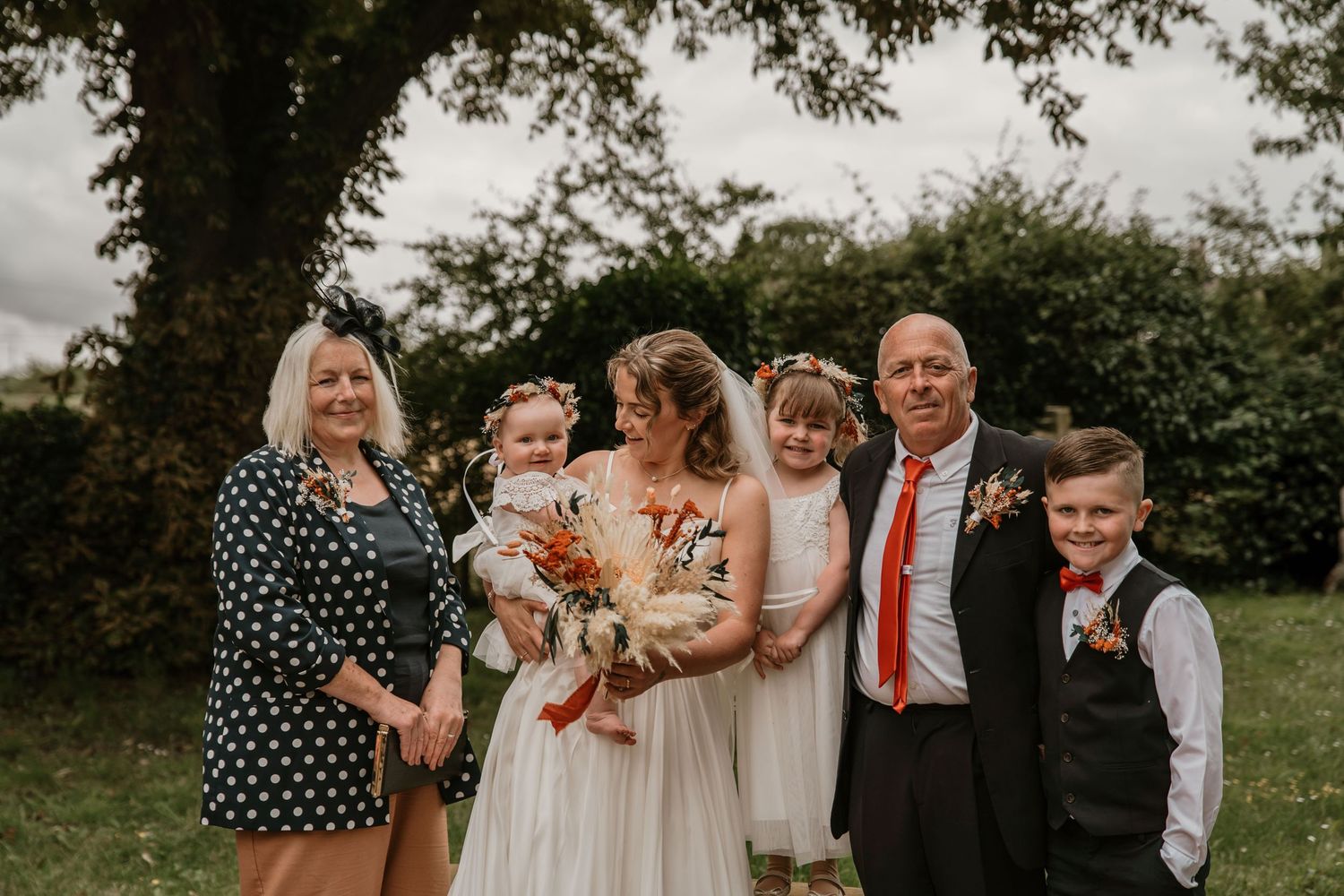 Bride and groom pose with family including children and grandparents in garden setting.
