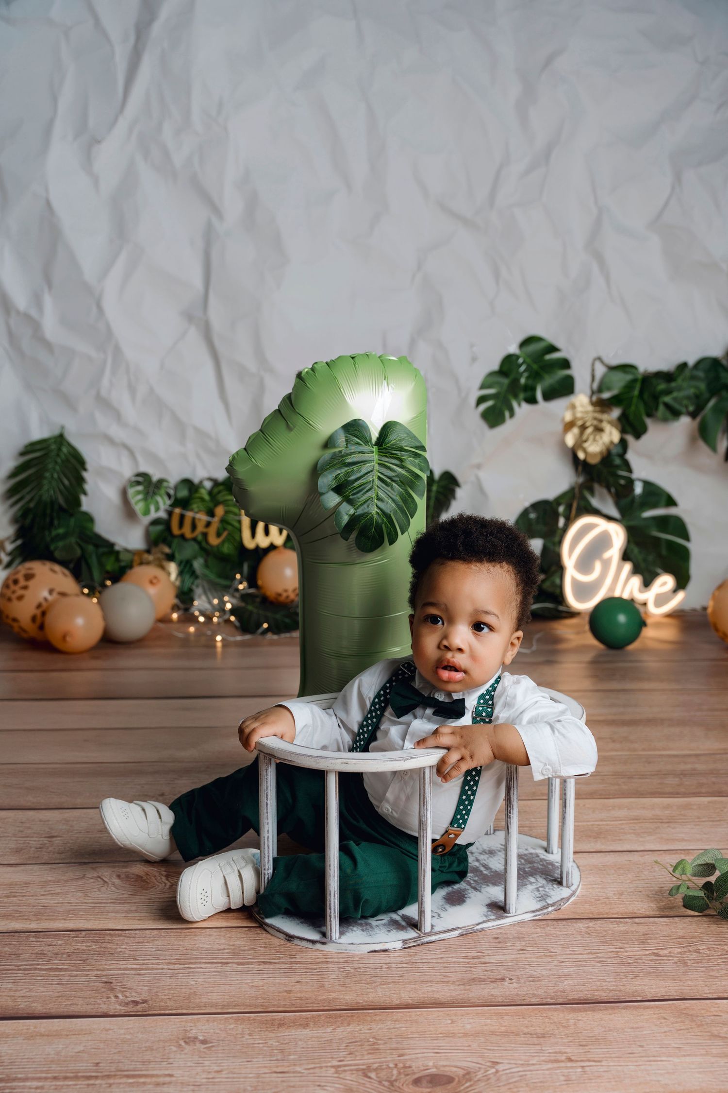 A toddler boy in green suspenders and bow tie sits on a wooden floor at a jungle-themed first birthday party.