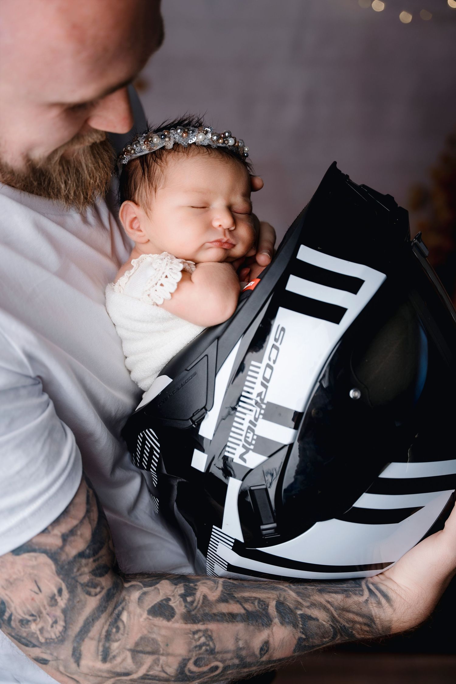 Tattooed father holding sleeping newborn baby girl in motorcycle helmet prop.