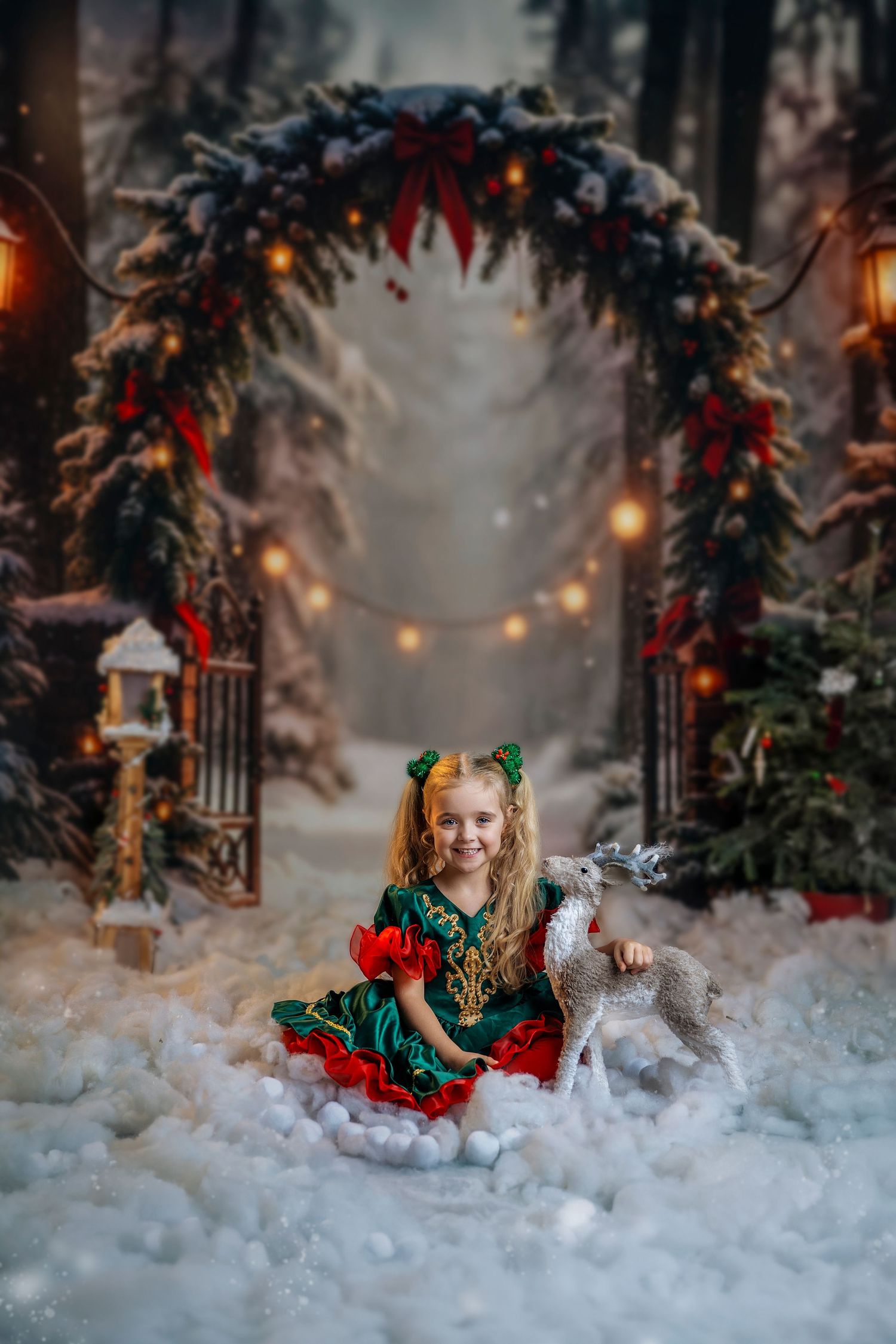 Young girl in green and red elf dress sits in snow beside reindeer figurine under festive Christmas arch with lights.