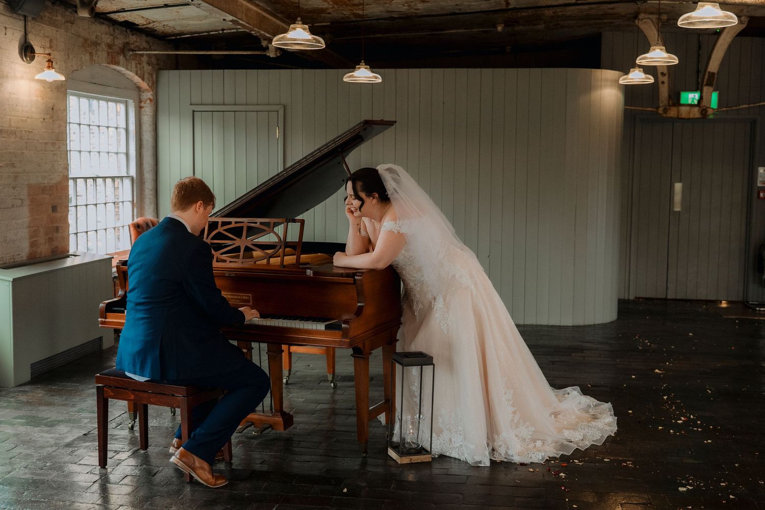 Bride and groom share romantic moment at grand piano in rustic industrial wedding venue.