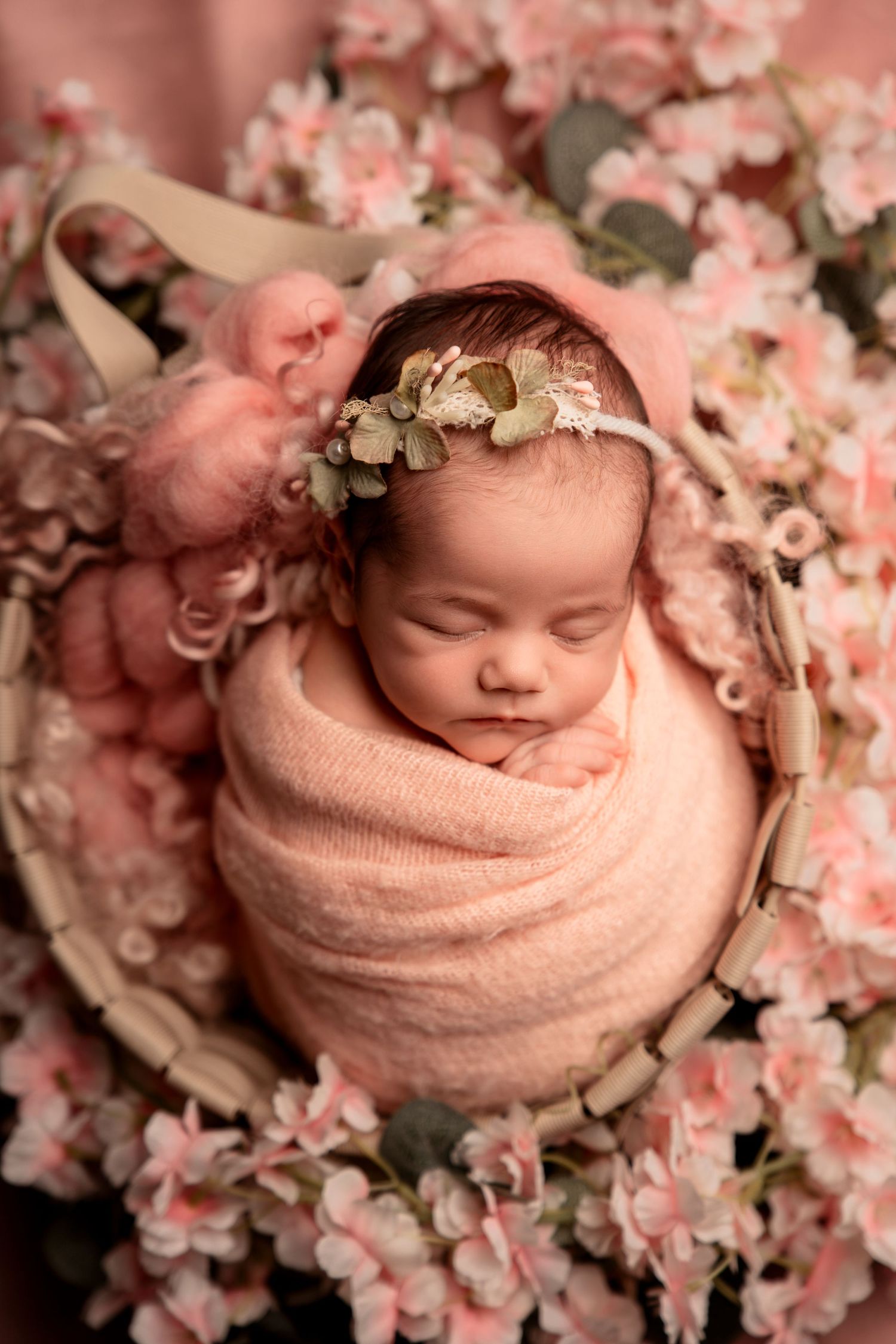 Newborn baby girl swaddled in pink wrap sleeping in a basket surrounded by pink flowers.