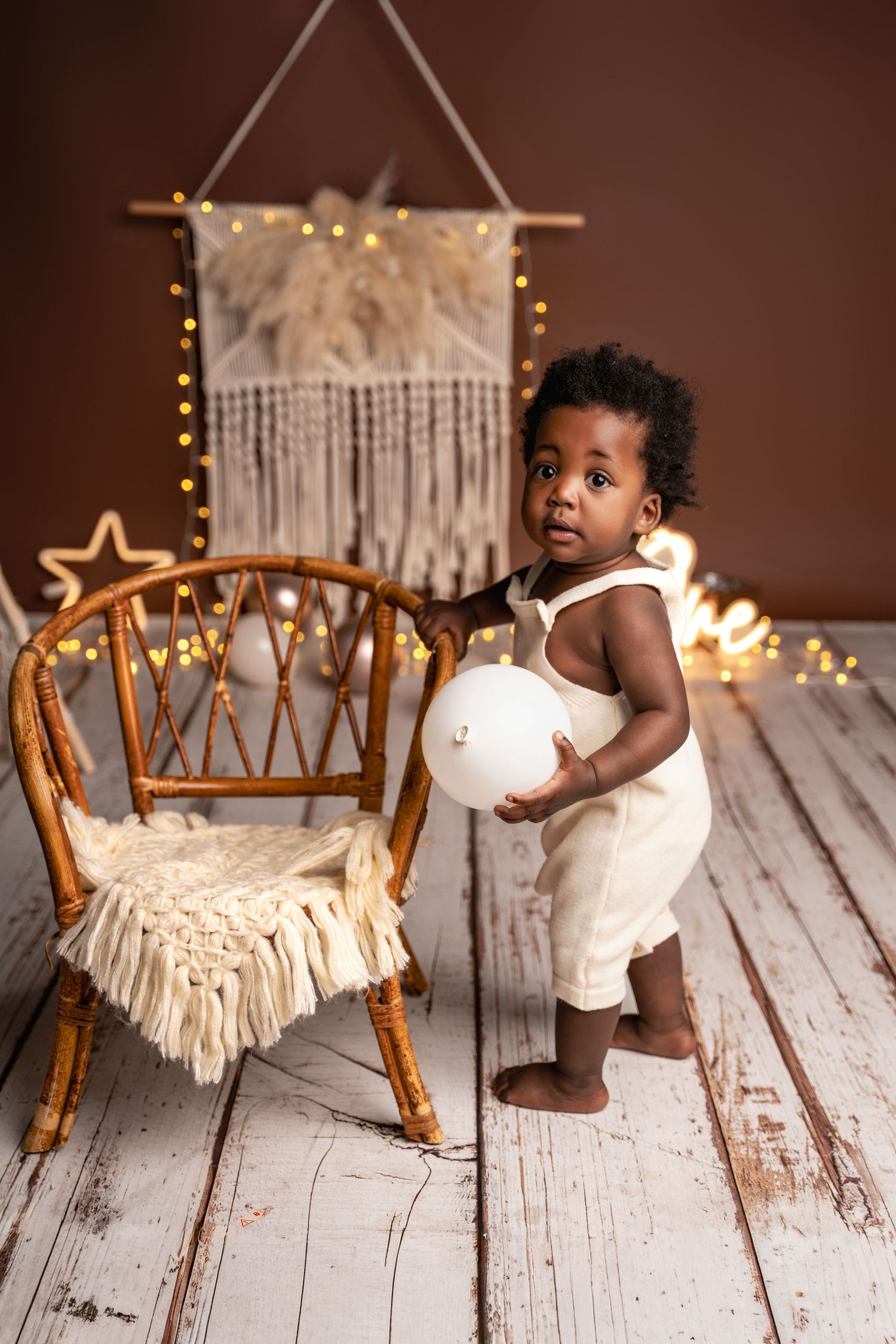 Adorable Black toddler in white romper holding balloon beside rattan chair with boho decor and fairy lights.