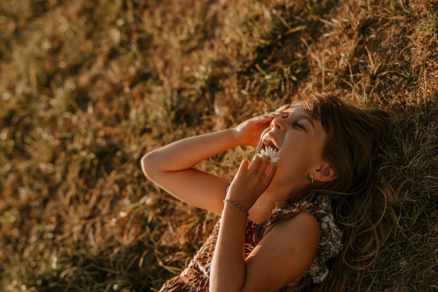 Young girl laughing joyfully while lying in golden dry grass field during warm sunset light.