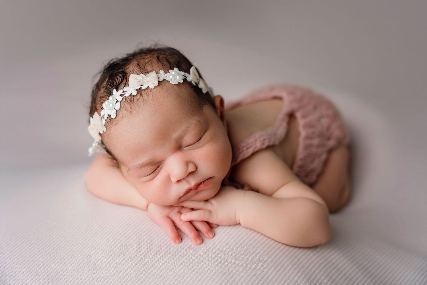 Sleeping newborn baby girl wearing a floral headband and pink knit outfit posed on a soft gray backdrop.