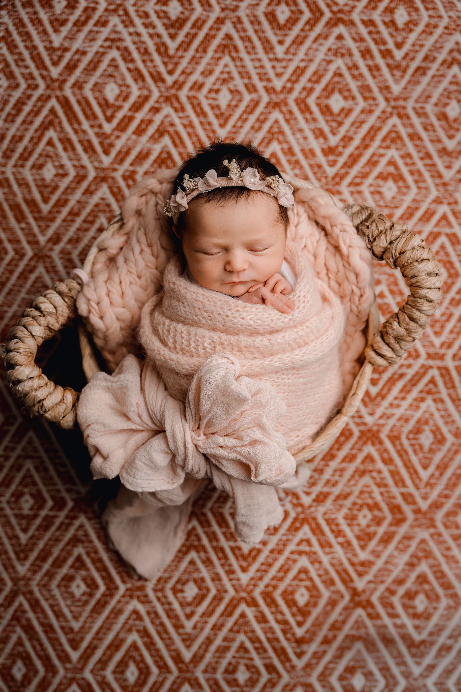 Sleeping newborn baby girl wrapped in pink knit swaddle with gold floral crown in a wicker basket.