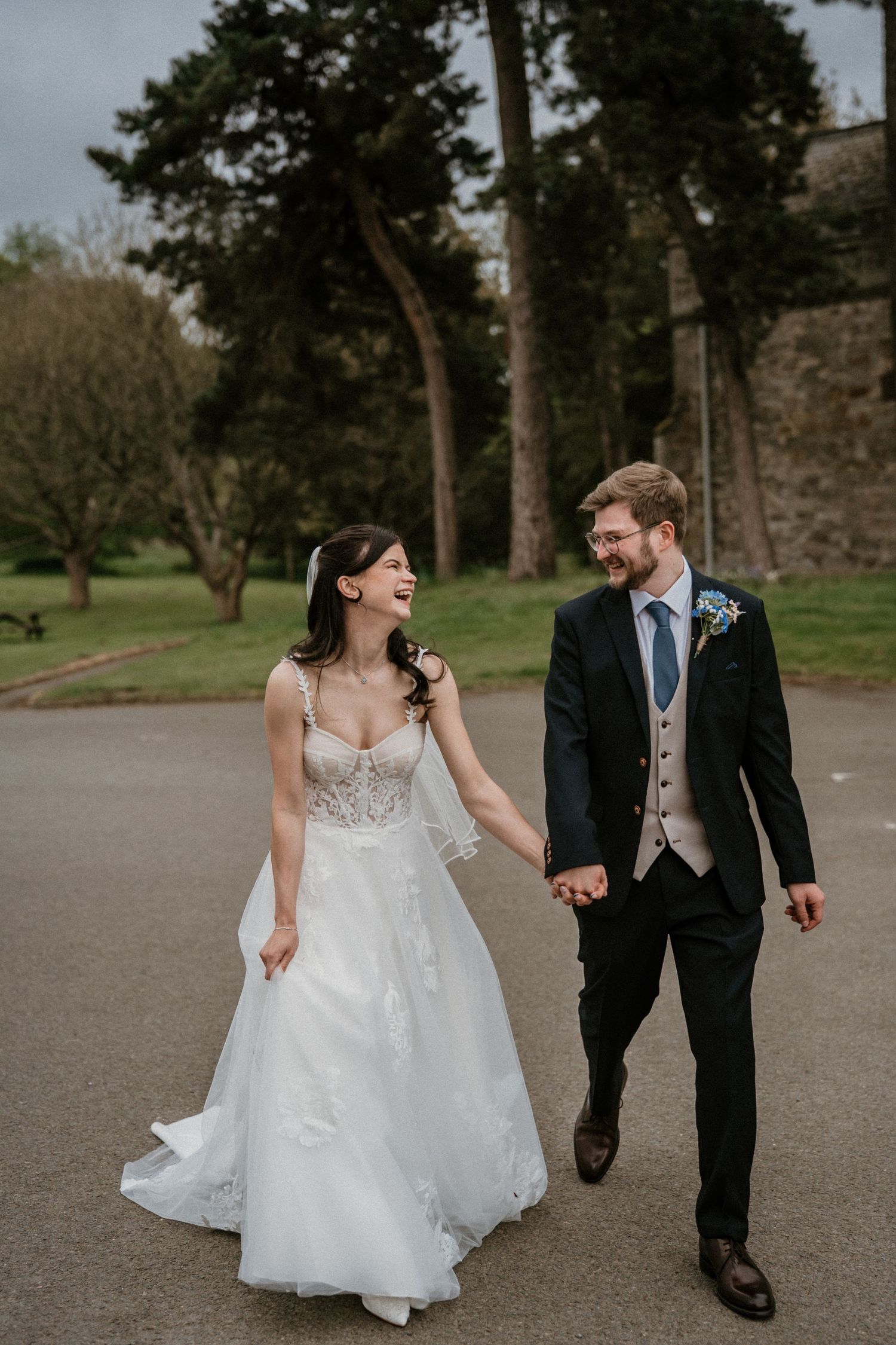 Laughing bride in lace gown holds hands with groom in navy suit walking outdoors on their wedding day.