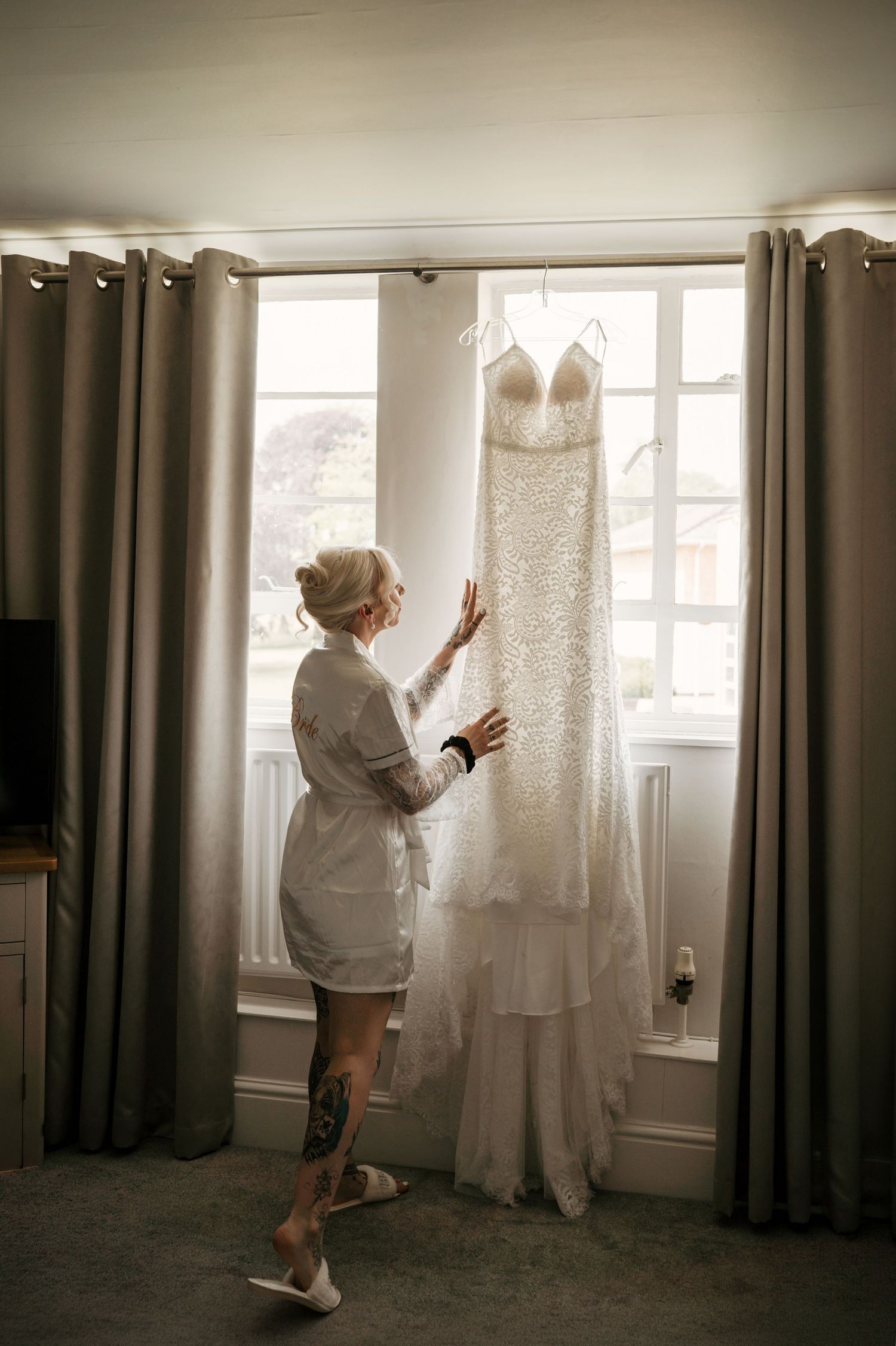 Tattooed bride in silk robe admires her lace wedding gown hanging by sunlit window before ceremony.
