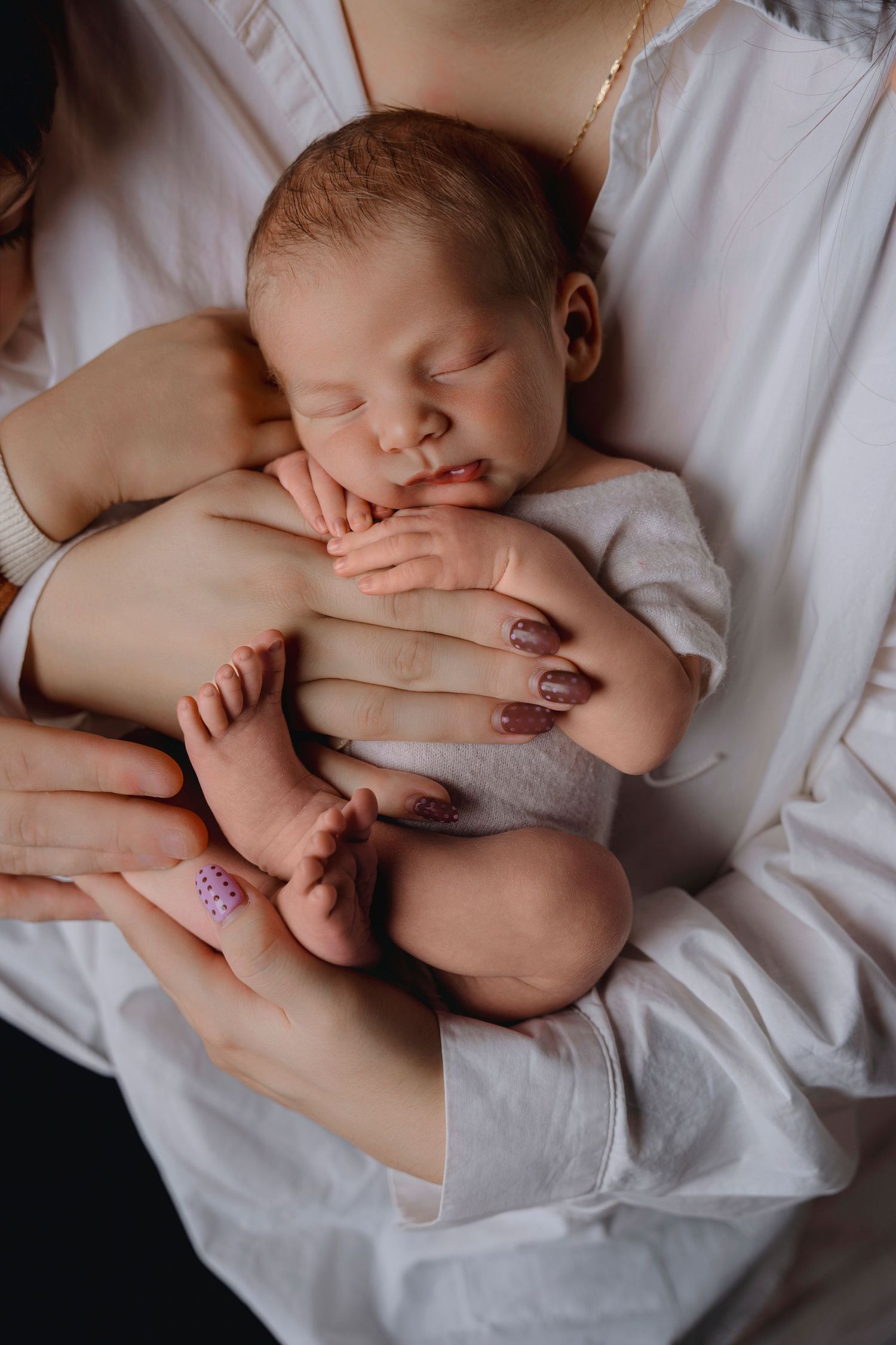 Mother in white dress cradling sleeping newborn baby with painted nails visible.