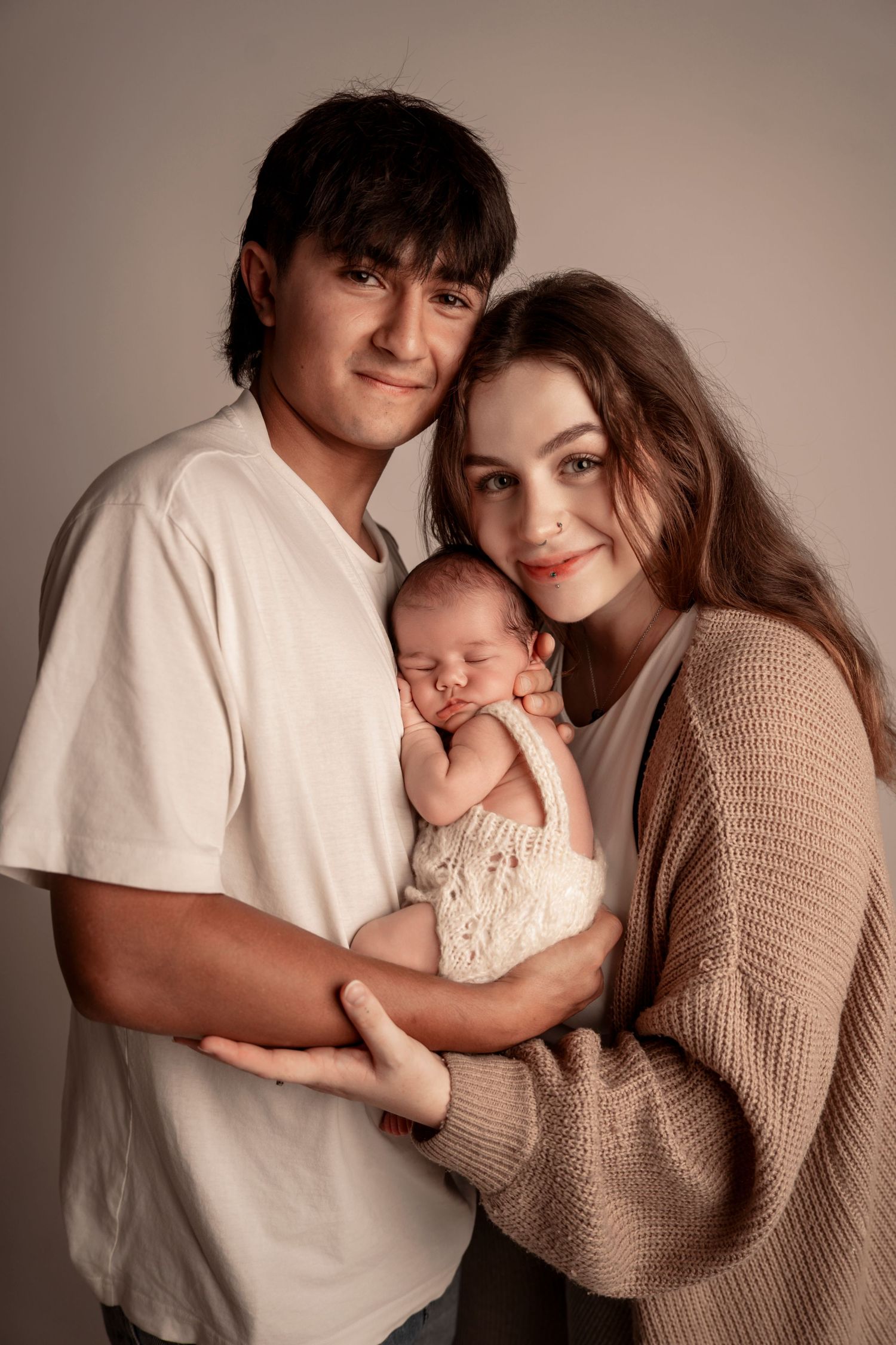 Young parents smiling and holding their sleeping newborn baby dressed in a cream knit romper.