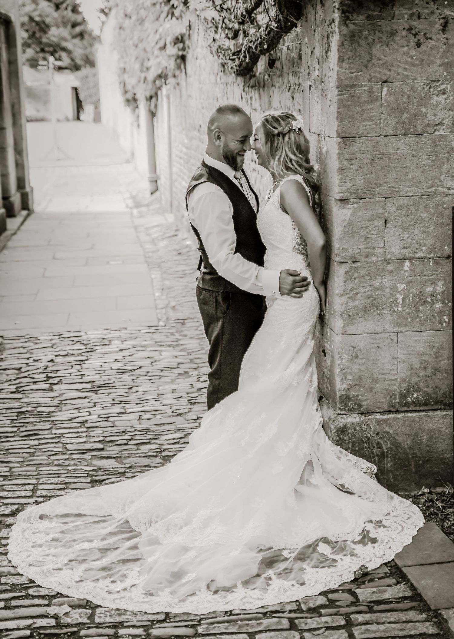 Black and white romantic portrait of bride and groom embracing against stone wall on cobblestone path.